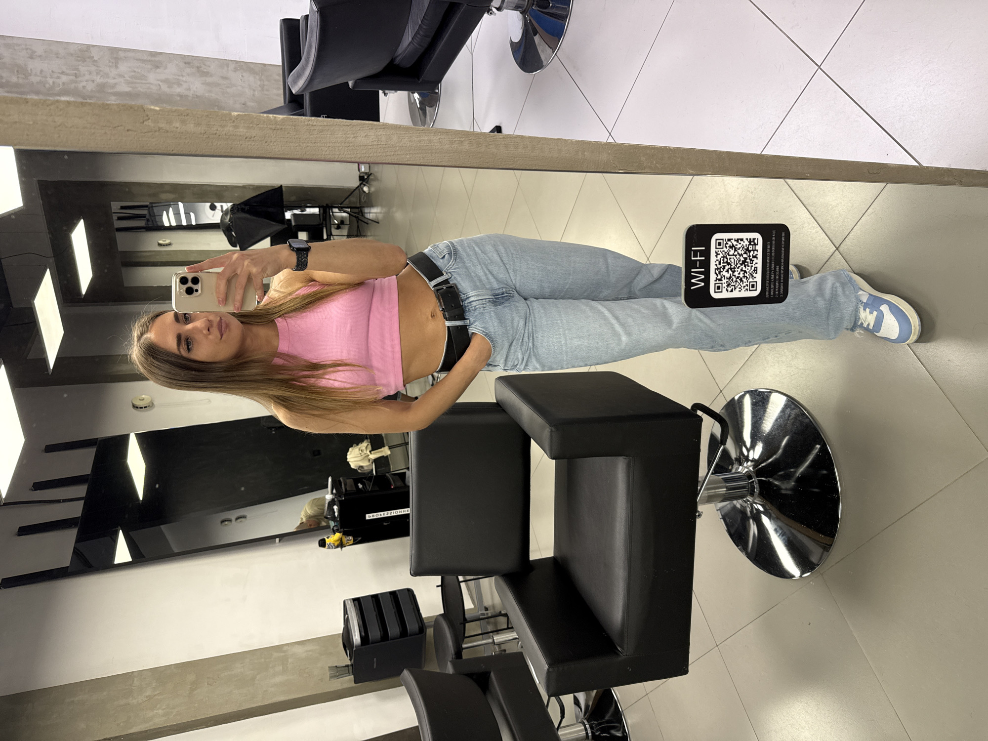 A young woman with long, straight, light brown hair is taking a selfie in front of a mirror inside what appears to be a hair salon. She is wearing a pink tank top and light blue jeans with a black belt. She is holding a mobile phone in her right hand, her face is visible. In the reflection of the mirror, there are several salon chairs, some black salon equipment, and lighting equipment. On the floor near her feet, there is a sign that says "WI-FI" with a QR code. The floor is tiled. The image appears to have been taken inside a salon, presumably located in Saint Petersburg, Russia, based on the Cyrillic text on the sign and the general aesthetics of the interior.
