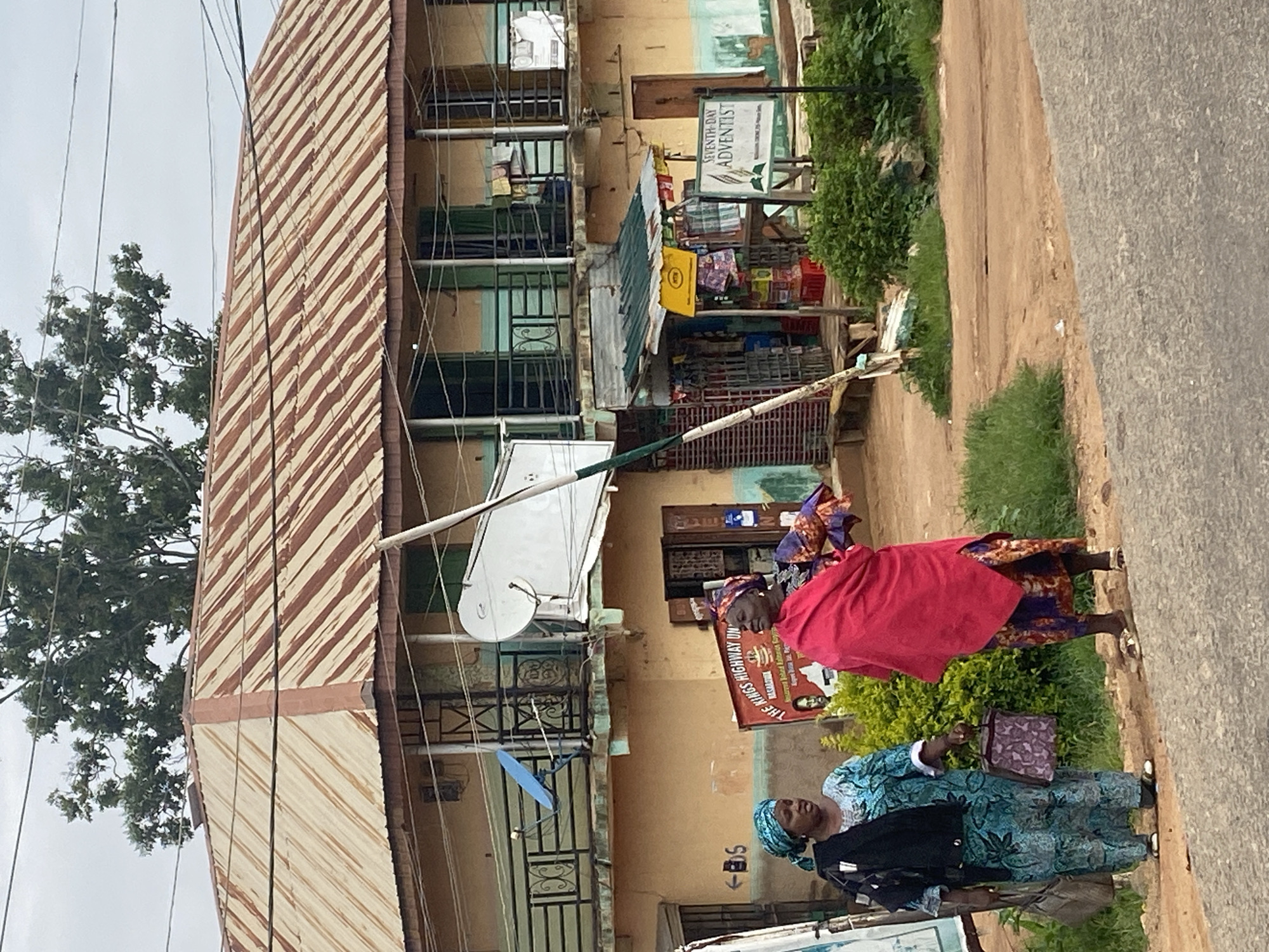 This is a medium shot, eye-level, outdoor, full-color photograph of a street scene in Jos, Nigeria, on an overcast day. The focal point is a two-story building with a corrugated metal roof that appears weathered and rusted. The building has balconies on both floors, with a black metal railing. A white satellite dish is mounted on the upper balcony. The walls of the building are painted a light yellow with a blue trim. On the ground floor, there is a small shop with goods displayed outside and a faded yellow sign. To the right of the shop, a white sign with green text reads "SEVENTH-DAY ADVENTIST". In front of the building, two women are standing on a dirt and asphalt road. The woman on the left is wearing a blue and green patterned dress and a blue headscarf, and is carrying two handbags. The woman on the right is wearing a vibrant red shawl over a purple and patterned dress, and a colorful headscarf. There is also a red and white sign visible behind the women, with text that is partially legible. Lush green vegetation is present on the right side of the image, contrasting with the earthy tones of the road and building. Electrical wires are visible crisscrossing the sky above the building.