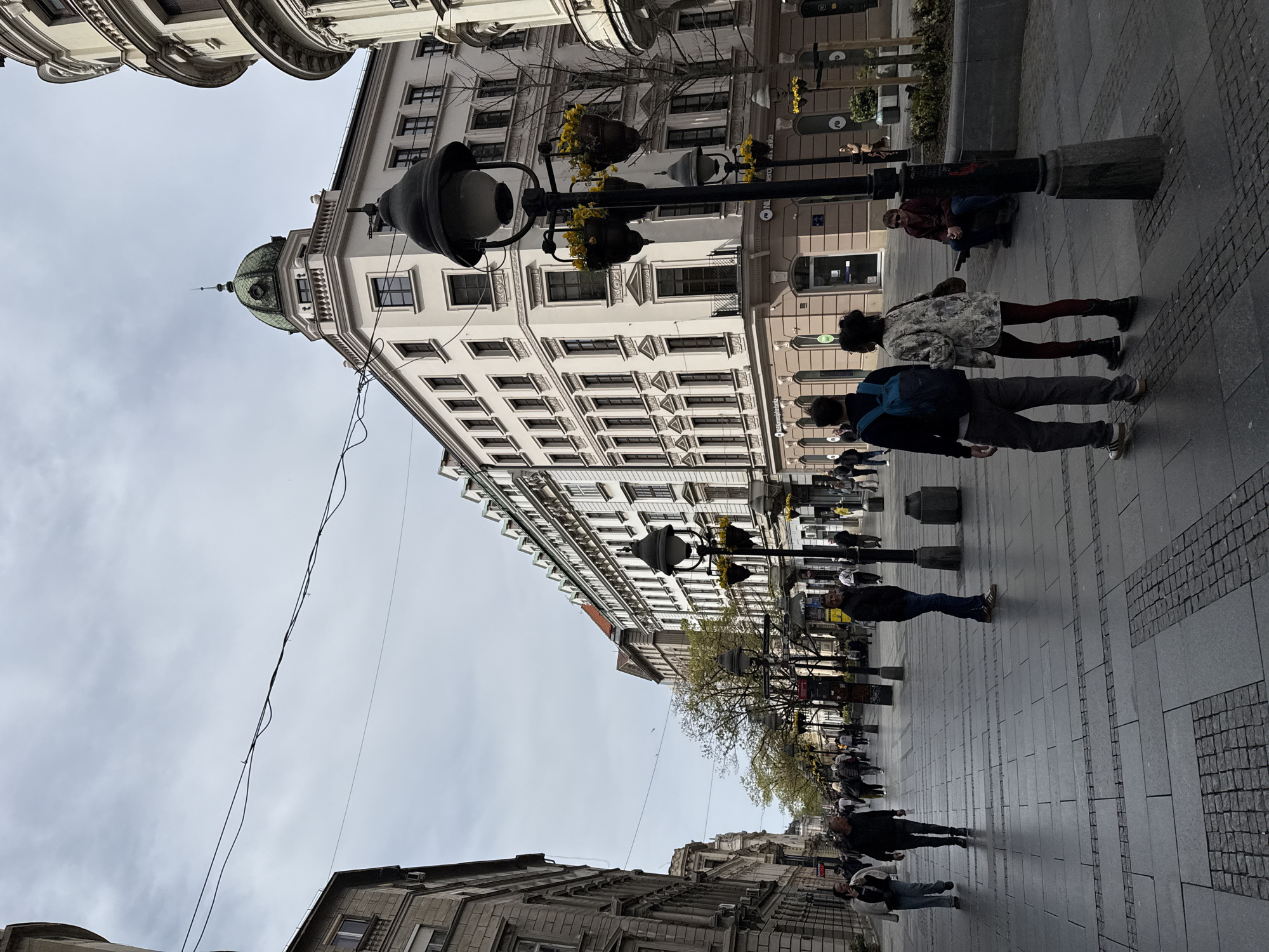 The image shows a street scene in Belgrade, Serbia. The perspective is looking up towards a building with a distinctive green dome on its corner. This likely identifies it as Knez Mihailova Street, a pedestrianized shopping zone known for its historical buildings. The street is paved with grey stones. Several people are walking along the pavement, including a couple walking together and others walking alone. A man is sitting on a bollard. There are ornate street lamps with hanging flower baskets positioned along the street. Wires run overhead. The sky is overcast. The buildings are ornate and multi-storied.