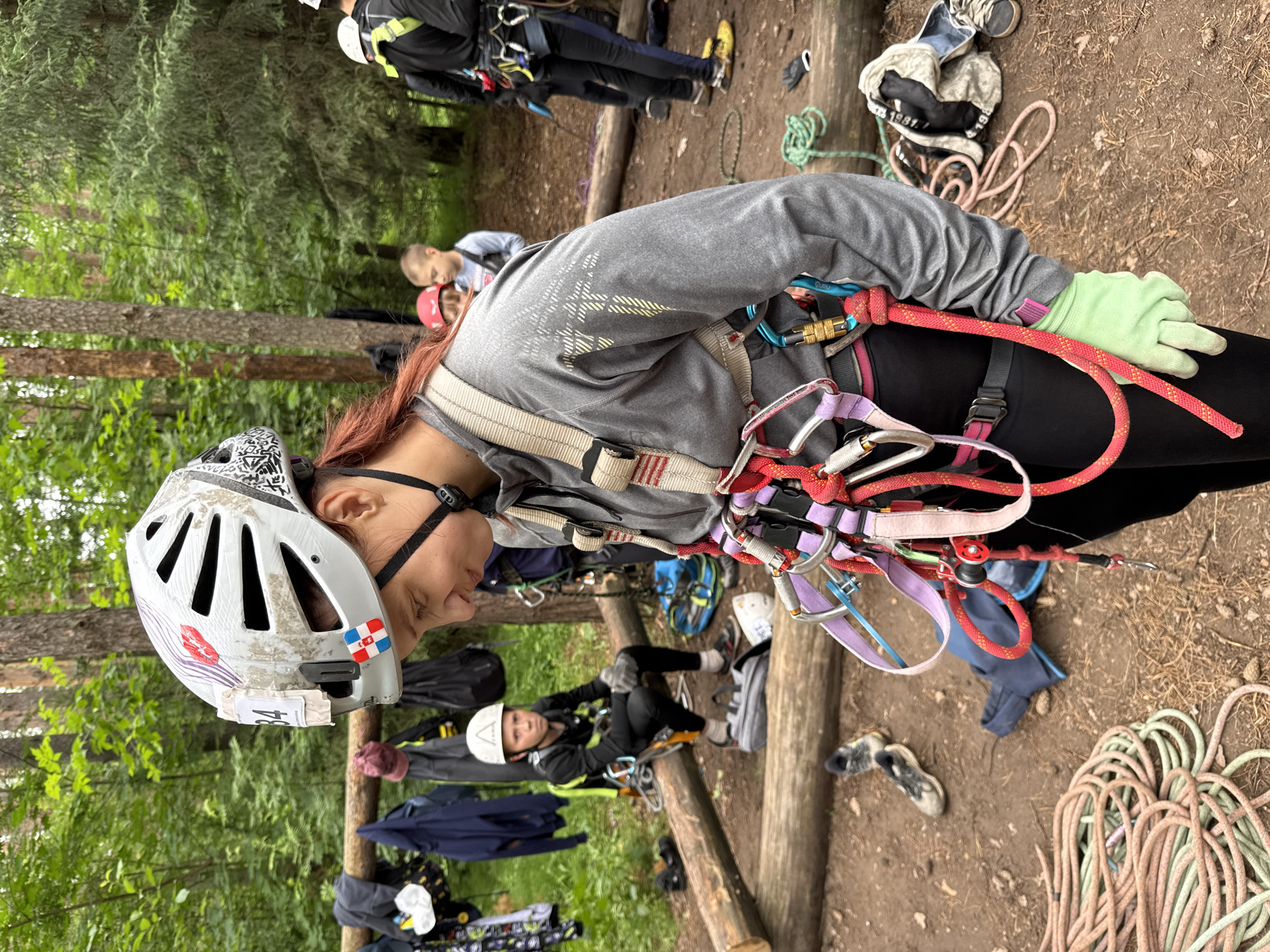 The image depicts an outdoor scene in a forest, likely an adventure park or training ground, potentially located in Chaikovsky, Russia, given the presence of a young boy wearing a helmet with a Russian flag emblem. In the foreground, a young woman with red hair is wearing a gray long-sleeved shirt, black pants, a climbing harness, and a white helmet with various stickers and a number "84" attached. She is looking down, and her harness is adorned with multiple carabiners, ropes, and a belay device.
Behind her, a young boy is sitting on a log, also wearing a helmet and harness, and appears to be participating in a climbing activity. Other individuals, dressed in athletic wear and safety gear, are visible in the background, with some standing and others in motion. The environment is characterized by numerous trees, logs used as seating or obstacles, and coiled ropes scattered on the ground. The overall atmosphere suggests an organized outdoor activity focused on climbing or adventure sports.