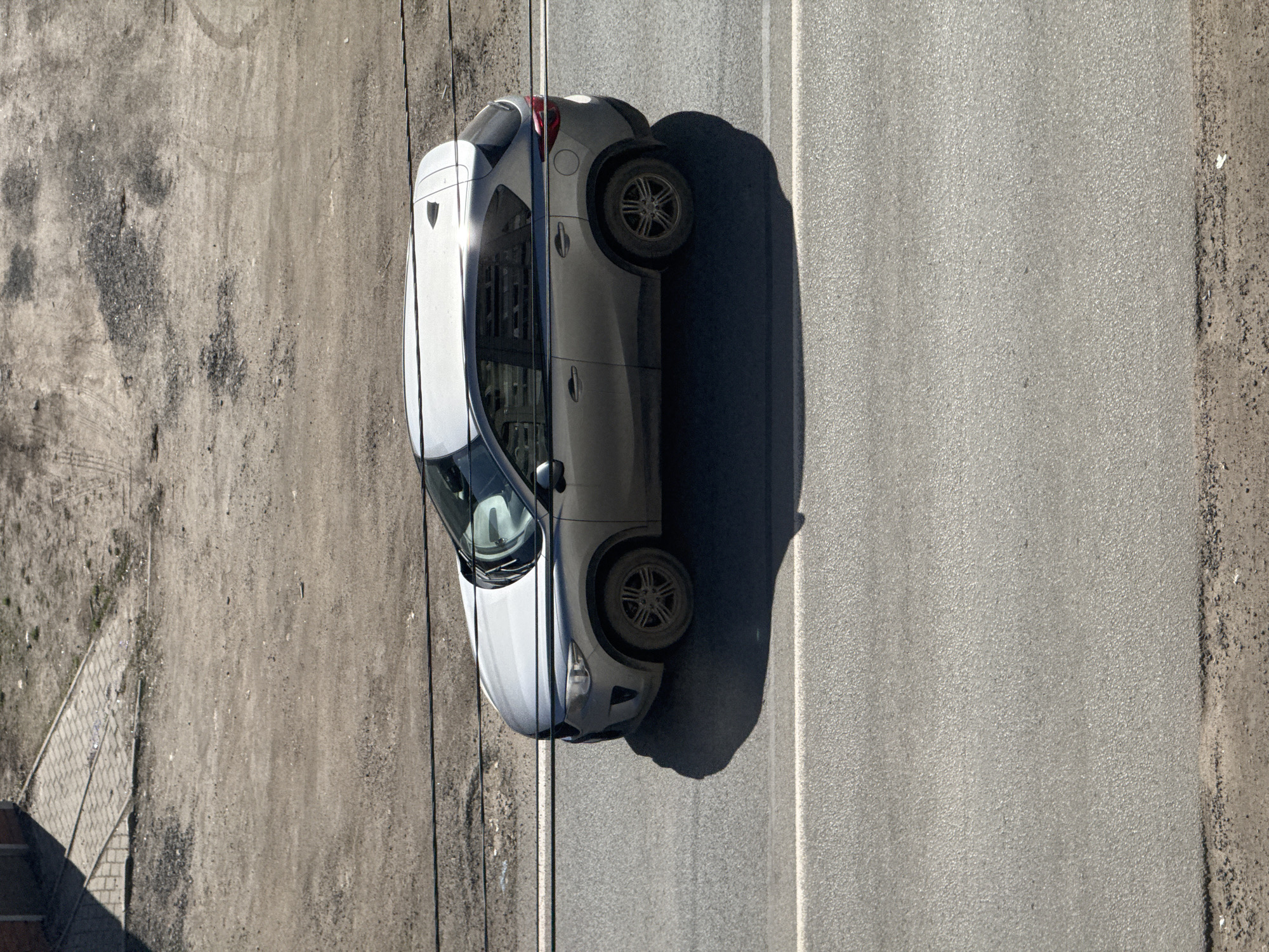 The image presents an overhead view of a light-colored SUV parked on the edge of a paved road next to an unpaved area. The perspective is looking straight down from a considerable height.
The main subject is a silver or light grey SUV. It is positioned vertically in the frame, with its right side on the asphalt road and its left side extending onto the rough, unpaved ground. Strong sunlight illuminates the scene, casting distinct, dark shadows of the car to its right and of thin, dark lines (likely overhead wires or cables) that cross the frame. Reflections of the sun are visible on the car's windshield and roof. The vehicle appears stationary.
The scene is divided into two main ground surfaces. The right side of the image features a paved asphalt road, gray and textured, with a continuous white line running along its left edge near the car. The left side is an unpaved, dusty, and somewhat barren dirt area, with scattered debris and small patches of ground cover. In the bottom-left corner, a section of a paved walkway made of interlocking bricks is visible, along with the corner of a dark structure casting a shadow.
The time of day is characterized by bright, direct sunlight, suggesting either mid-morning or afternoon. The weather is clear and sunny. The location cues include a typical road, an undeveloped dirt shoulder, and visible infrastructure like wires and a paved path. There is no discernible text in the image. The setting is an outdoor area in Ново-Никольское, Russia.