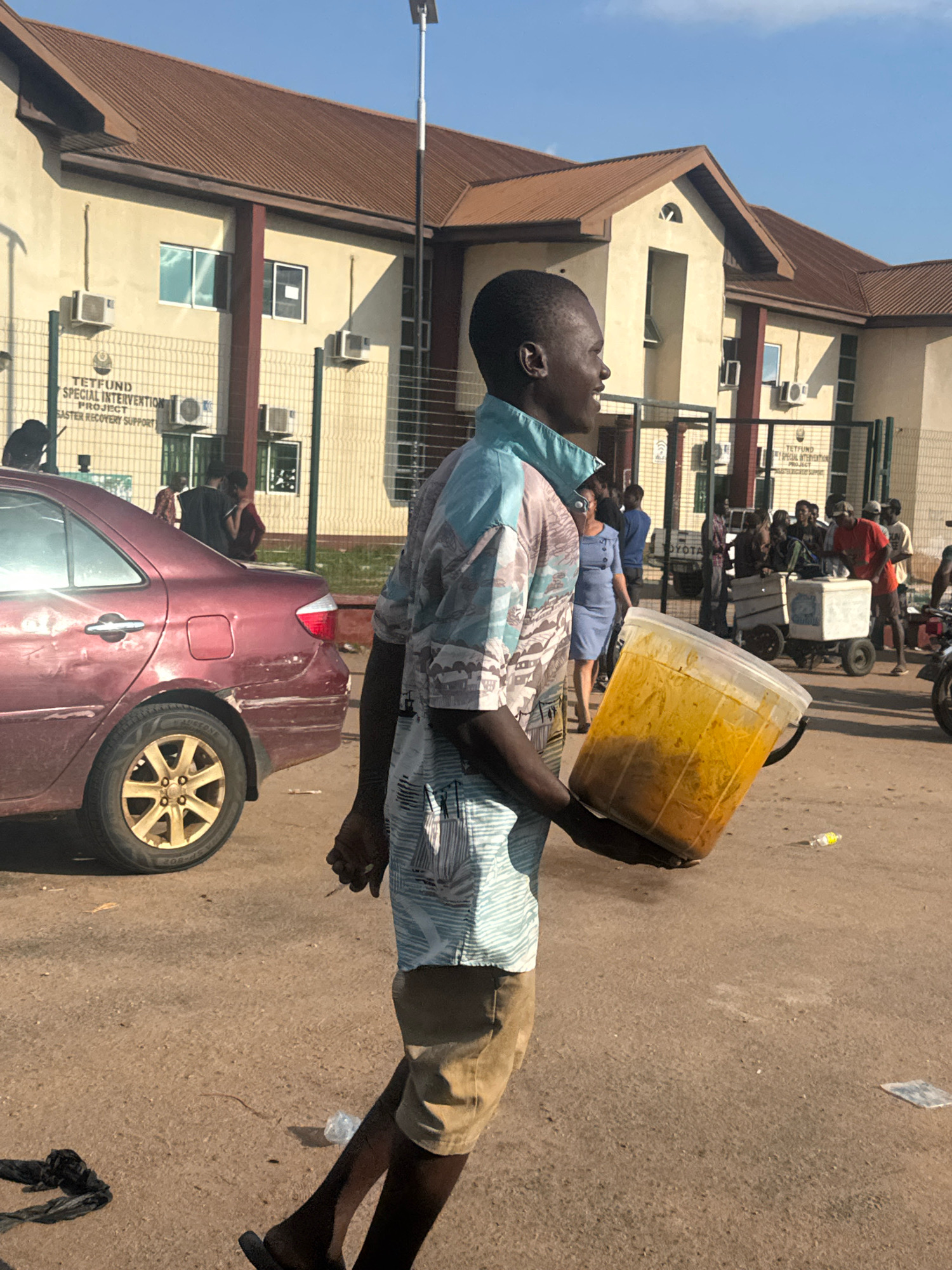 In Ihiagwa, Nigeria, a young man walks across a dusty street carrying a large, clear plastic container filled with a yellow, stew-like substance. He is wearing a short-sleeved, patterned shirt and light-colored shorts. Behind him, a maroon sedan is parked, its rear tire and bumper visibly damaged. A building with a brown roof and beige walls stands in the background, with air conditioning units mounted on its exterior. A metal fence encloses part of the building, with a sign displaying text that reads "TETFUND SPECIAL INTERVENTION PROJECT DISASTER RECOVERY SUPPORT." Other people are visible in the background, some interacting with each other and others near a white cooler on wheels. The scene is under a clear blue sky with sunlight casting shadows.