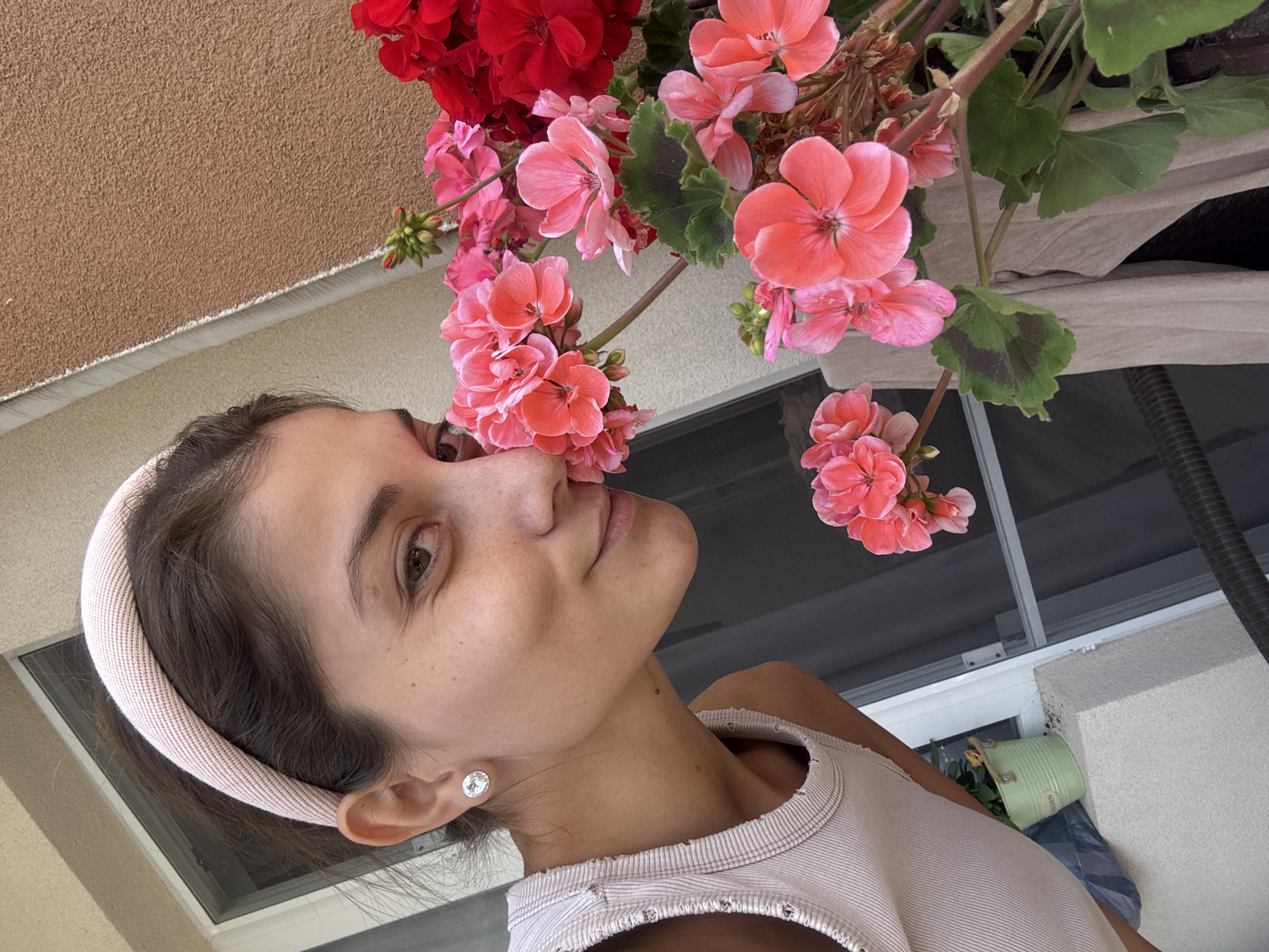 A young woman poses with a blooming flower pot, her face turned upwards towards the flowers. She wears a light-colored tank top and a matching headband. A stud earring is visible in her left ear. The flowers are a mix of red and pink geraniums. Behind her, a window and balcony area are visible. The location is not identifiable as Burgas, Bulgaria from the provided image.