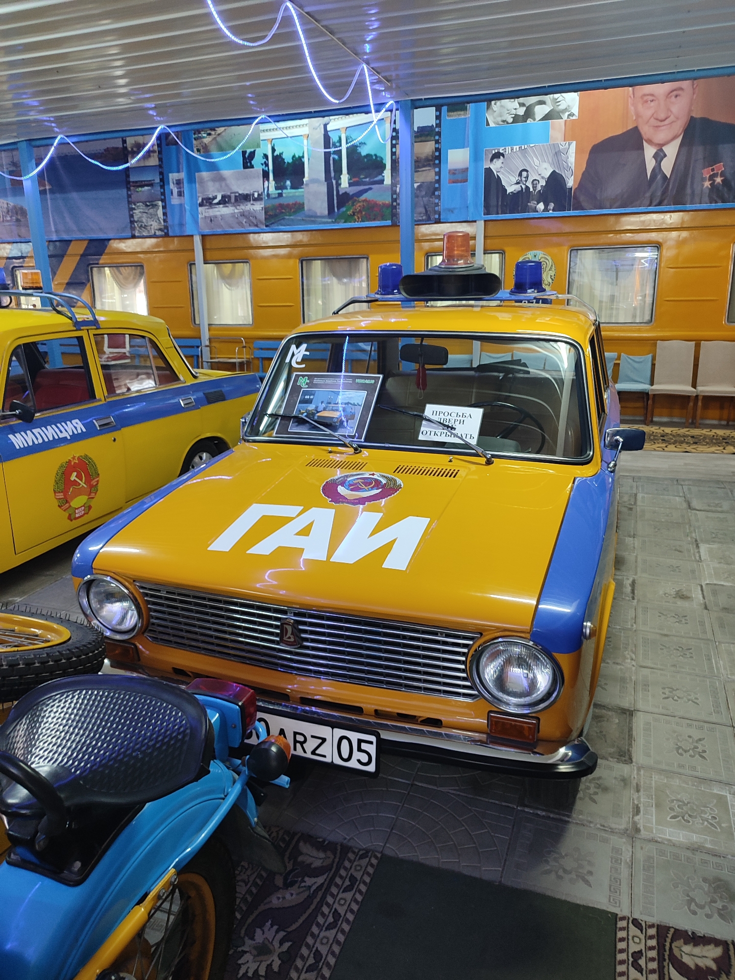 In the city of Qonaev, Kazakhstan, an indoor display features vintage Soviet-era vehicles. The central subject is a yellow and blue car, likely a Lada, marked with the acronym "ГАИ" (GAI) in large white letters on its hood, indicating it was a traffic police vehicle. It has a blue stripe along its side and a blue roof with orange and blue emergency lights. The car's windshield displays a sign reading "ПРОСЬБА ДВЕРИ ОТКРЫТЬ" (PLEASE OPEN DOORS). To the left of this car is another yellow car, also bearing a blue stripe and the word "МИЛИЦИЯ" (MILITSIYA - police) on its side, along with a Soviet emblem. The background consists of a large mural depicting Soviet-era scenes and portraits, including one of a man in a suit and medals. A blue motorcycle with a black seat is partially visible in the foreground, to the left of the yellow and blue car. The vehicles are displayed on a tiled floor, with a patterned rug partially visible. The overall environment suggests a museum or exhibition space.