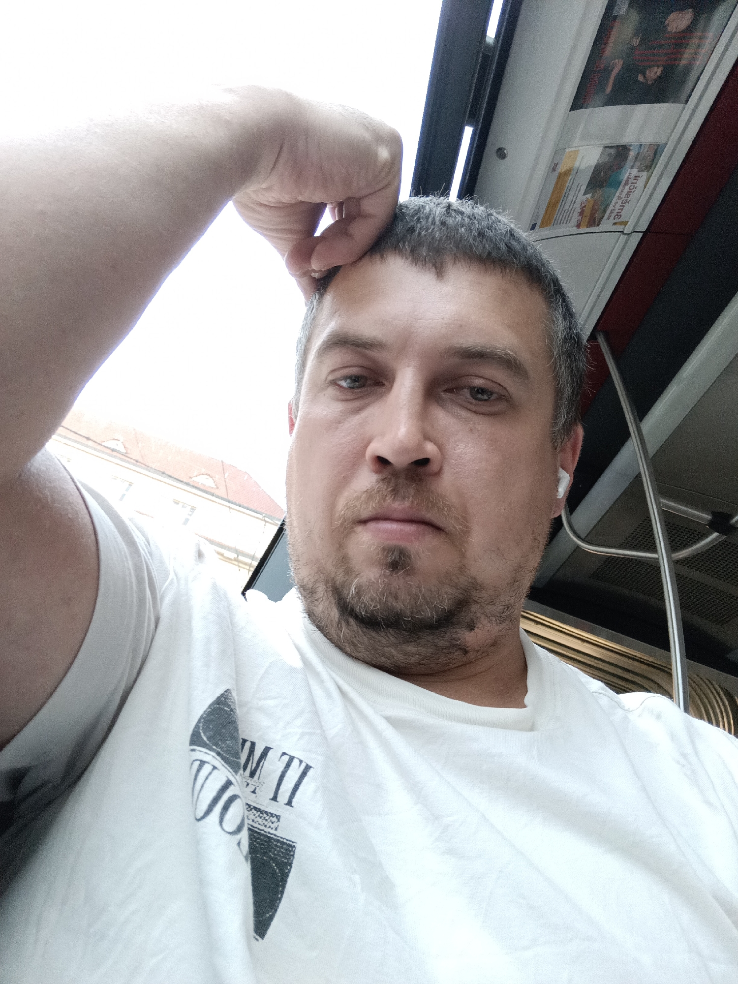 A close-up, selfie-style photograph shows a middle-aged man with short, graying hair and a goatee, wearing a white t-shirt. He has his left arm raised and resting on his forehead, and he is wearing a white wireless earbud in his right ear. The man appears to be inside a public transport vehicle, likely a bus or tram, as suggested by the metal poles and advertisements visible in the background. The man's expression is neutral, and he is looking directly at the camera. The lighting suggests it is daytime, possibly with sunlight filtering in from outside. The background details, such as the style of the advertisements and the interior design of the vehicle, are consistent with public transportation found in Prague, Czechia.