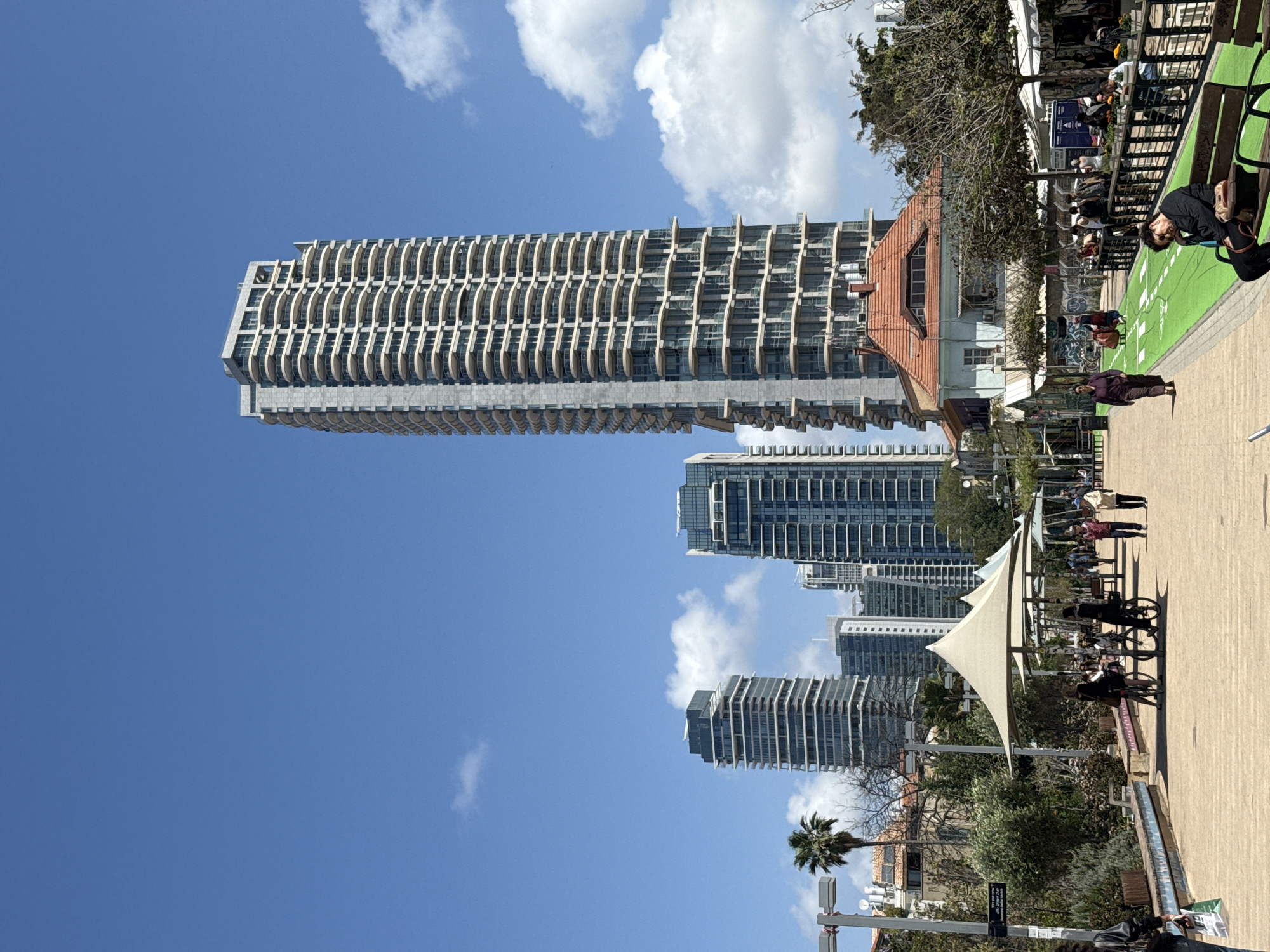 This image captures a bustling urban scene in Tel-Aviv, Israel, under a clear blue sky dotted with fluffy white clouds, suggesting a bright and sunny day. The dominant architectural feature is a striking, very tall modern skyscraper on the left, characterized by its numerous curved, ribbed balconies that create a distinctive, wave-like facade. Several other contemporary high-rise buildings with glass exteriors are visible further back and to the right, adding to the city's modern skyline.
In stark contrast, a smaller, older building with a red-tiled roof and pale walls stands in the middle ground, surrounded by trees. On the right, a wide, paved promenade is alive with activity. People are seen enjoying the outdoor space; some are walking, while others are seated at tables under white awnings, indicative of cafes or restaurants. One person in a dark top sits on a long, green bench-like structure, which runs alongside a green-colored strip on the pavement, possibly a dedicated bike lane or pathway. Another individual is seen walking away from the camera.
Lush green trees and a prominent palm tree are scattered throughout the scene, providing natural elements against the urban backdrop. The strong sunlight and clear skies suggest it is midday or early afternoon. A small sign on a pole in the lower right corner contains illegible text, likely in Hebrew, given the location. The overall atmosphere is lively and inviting, showcasing a blend of modern and traditional elements in a vibrant city setting.