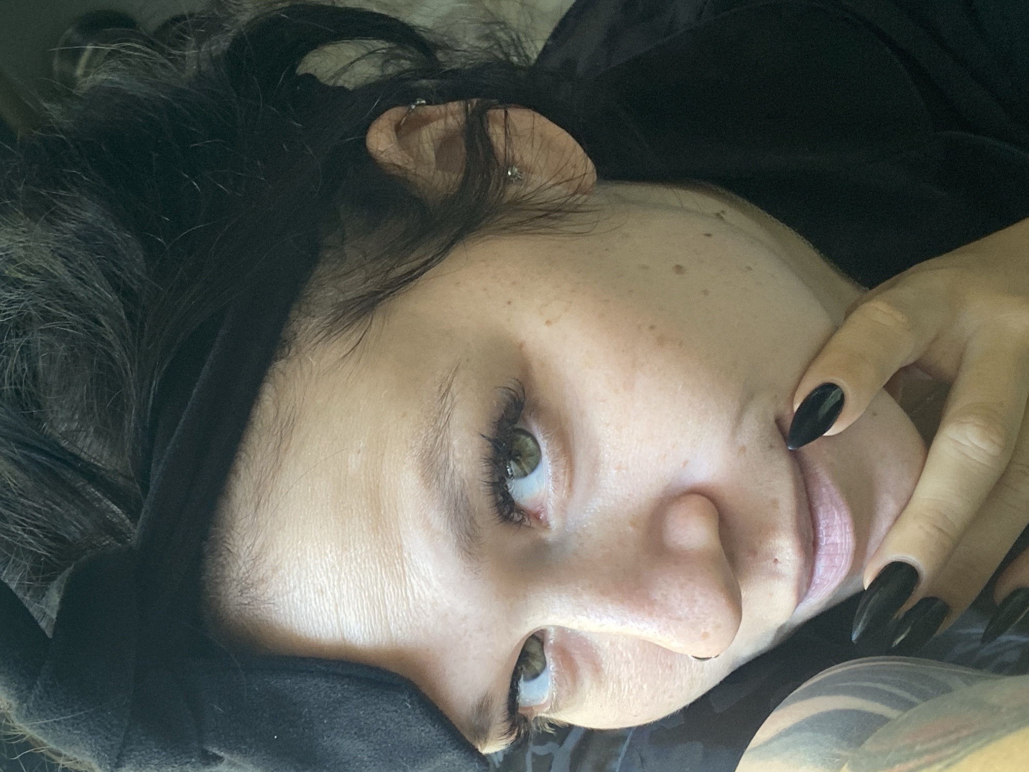 This is a close-up, medium shot, full-face portrait of a woman. She has dark hair pulled back with a black headband that resembles a bow. Her eyes are green and she is looking towards the left side of the frame. She has long, dark eyelashes and appears to be wearing makeup. She has a nose ring on her left nostril. Her skin has freckles and blemishes. She is wearing a black top. Her left hand is resting on her chin, and her fingernails are painted black and are long and pointed. The background is dark and out of focus, making the woman the central subject. There are no discernible elements in the image that would indicate a specific location in Chaikovsky, Russia.