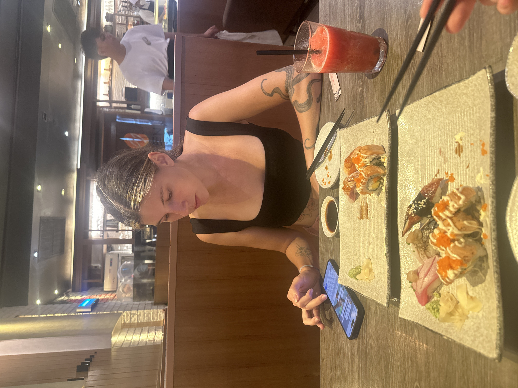 Here is a detailed description of the image:
A woman with tattoos on her arm sits at a wooden table in a restaurant. She is wearing a black tank top and looking at her phone. Two plates of sushi are on the table, along with a drink, a small dish of soy sauce, and chopsticks. In the background, a man in a white shirt stands behind a counter. The setting appears to be a restaurant, potentially in or near บ้านบางรักษ์, Thailand based on the location. The woman seems to be engaged with her phone while having a meal at the sushi restaurant.