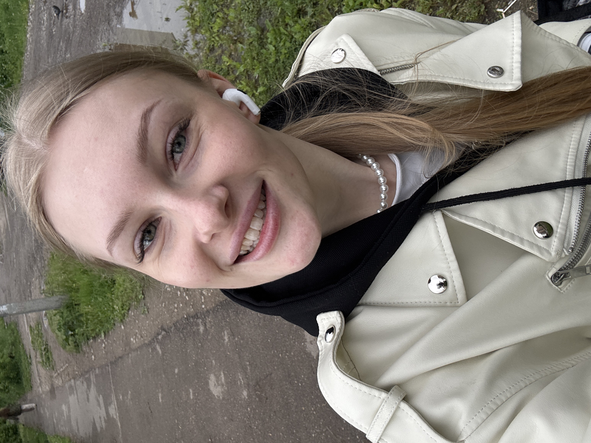 The image captures a close-up selfie of a young woman outdoors in Perm, Russia. She has light blonde hair, green eyes, and fair skin, with a clear complexion. She is smiling broadly, revealing her upper and lower teeth, and looking directly into the camera. She wears a white motorcycle-style jacket, partially open, over a black hooded sweatshirt and a white t-shirt. A single-strand pearl necklace is visible around her neck, and she has white wireless earbuds in both ears. Her hair is pulled back in a low style, with some loose strands falling over her right shoulder.
The background shows a worn, grey asphalt path with several puddles reflecting the sky. Green grass and low vegetation border both sides of the path. In the far distance on the path, a single person is walking away from the camera. A grey utility pole stands upright to the right of the path in the mid-ground. A small, dark bird is visible on the path near a puddle in the upper right portion of the background. The lighting is diffused, suggesting an overcast day.