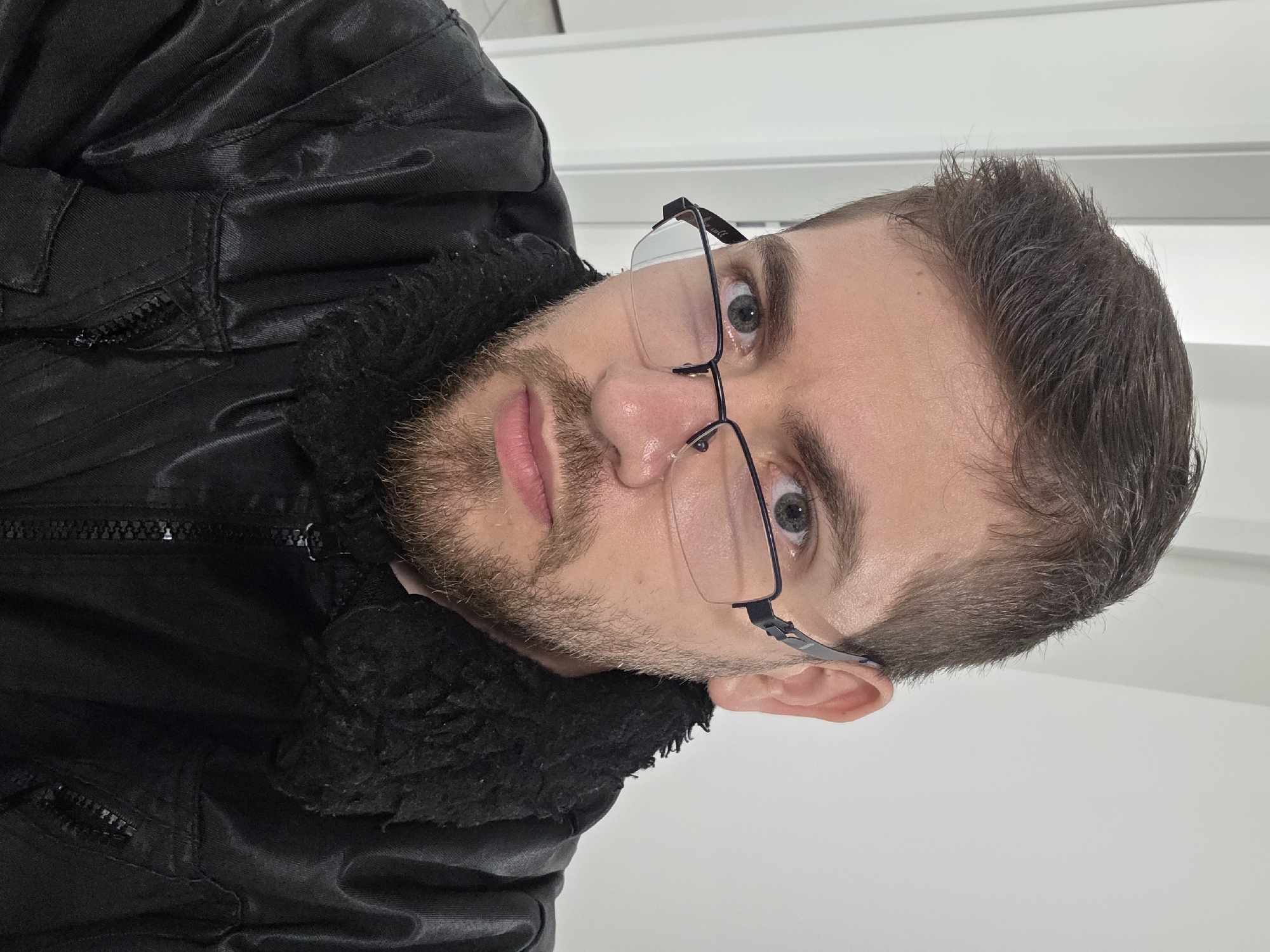 A man is positioned close to the camera, capturing a head and upper torso shot. He is facing the camera at a slight angle and is wearing eyeglasses with thin black frames. His facial hair consists of a stubble beard and mustache. He is wearing a dark jacket with a faux fur collar, and a partial zipper is visible on the left side of the frame. The background is a plain, light-colored wall. The photo appears to be taken indoors under bright, artificial lighting. The location is unspecified.