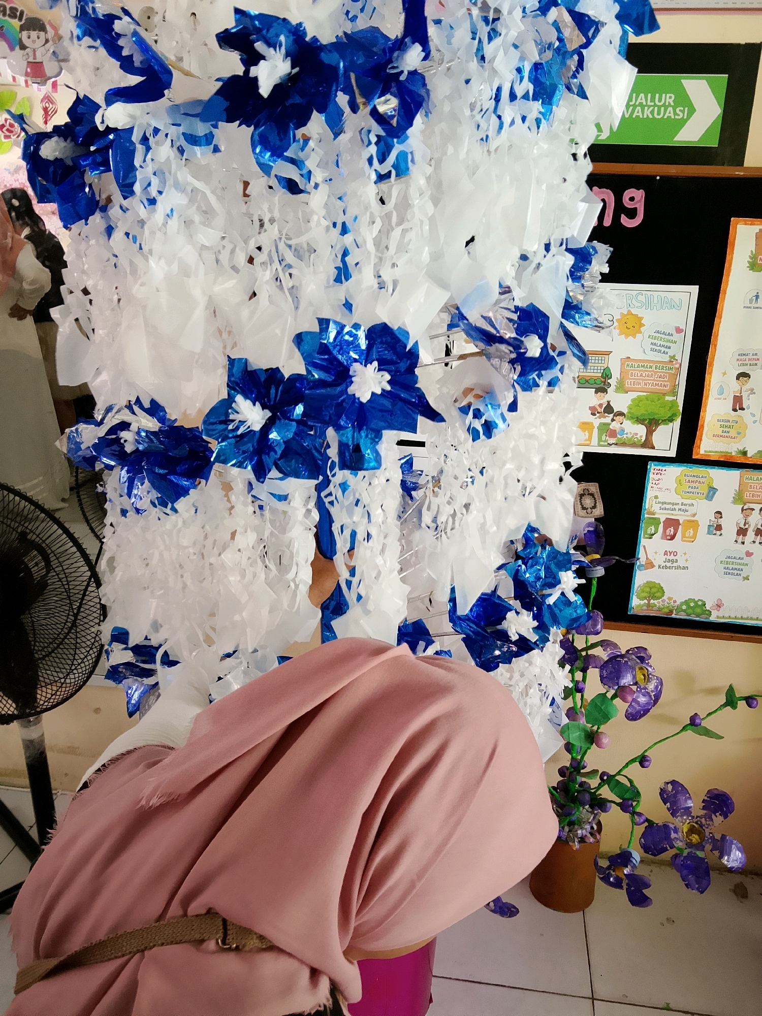 A person wearing a pink hijab and a brown woven belt is partially visible in the foreground, their head obscured by the hijab. They are facing away from the camera, with their back towards a large, elaborate decoration made of white and blue paper streamers and metallic blue flowers. To the left of the decoration, a black oscillating fan is visible, and to the right, a potted artificial plant with purple flowers and green leaves stands on a tiled floor. In the background, a blackboard is mounted on a wall, with a green sign that reads "JALUR EVAKUASI" (Evacuation Route) in white lettering and an arrow pointing to the right. Other posters are also visible on the wall, including one with the text "JAGALAH KEBERSIHAN HALAMAN SEKOLAH" (Maintain the cleanliness of the school grounds) and another with an illustration of children and trees. The scene appears to be indoors, possibly in a school setting in Besusu Timur, Indonesia.