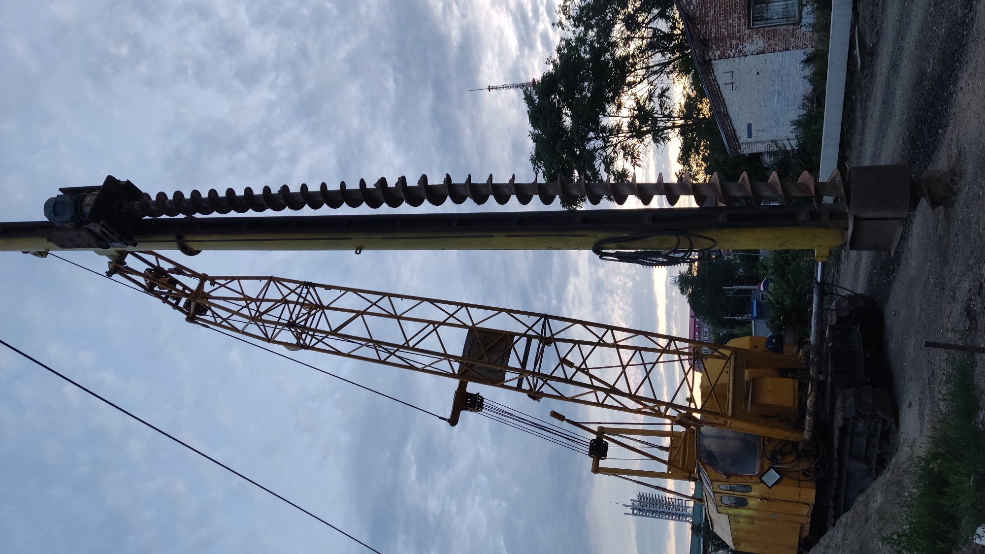 A large yellow crane, equipped with a drill auger, is positioned on a construction site. The crane's boom is angled upwards and to the left, with its heavy machinery visible at the base. The auger, a long, spiral drill bit, is suspended vertically next to the crane. The sky is overcast with a few patches of lighter clouds, suggesting either early morning or late evening. In the background, trees and some buildings, including a brick structure with a white painted section, are visible. A tall radio tower or antenna can be seen in the distance. The ground appears to be a mixture of dirt and gravel. The overall scene depicts an active construction or engineering project.