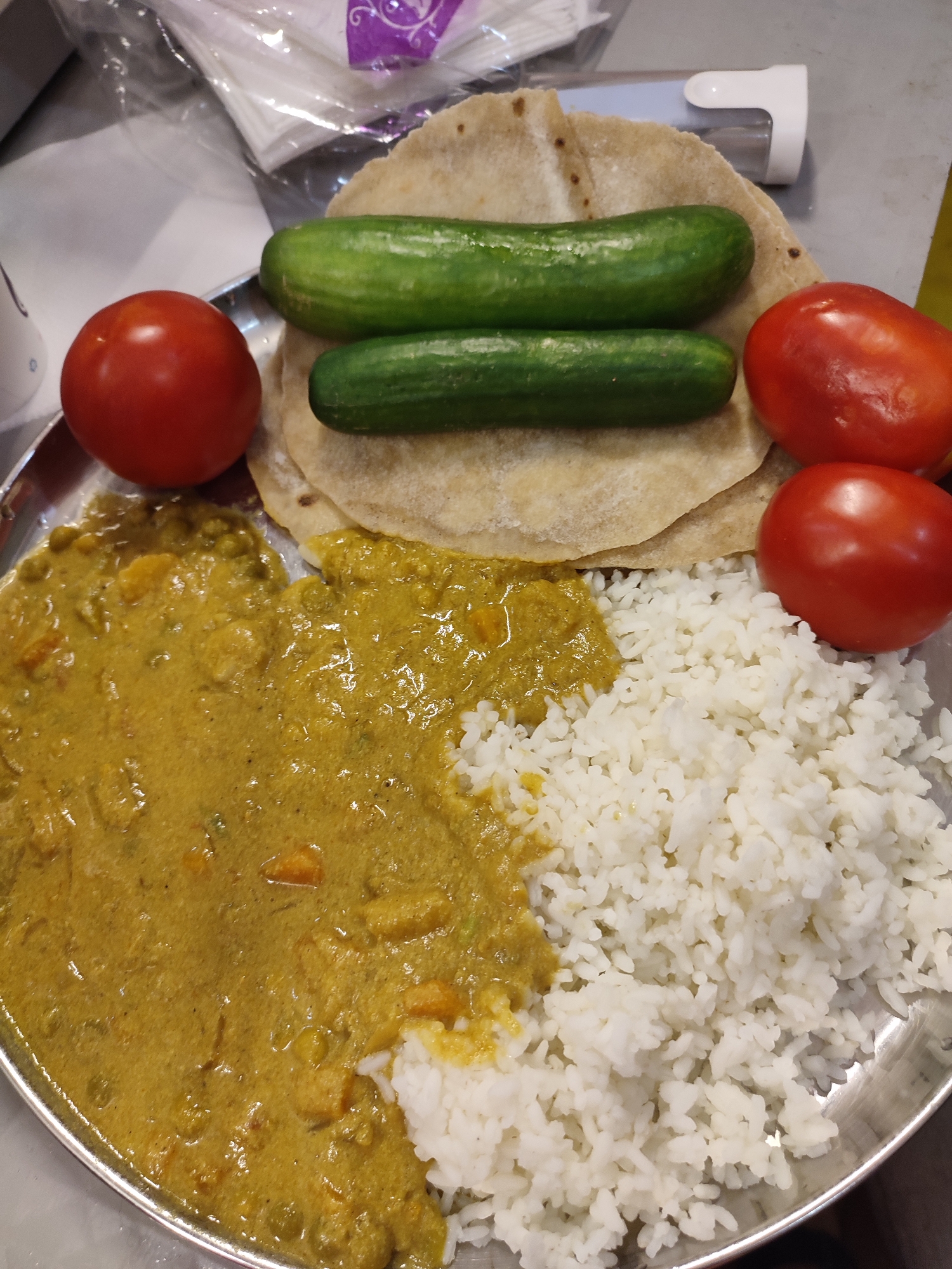This image presents a simple meal served on a round, metallic plate, common in Indian households or eateries. The plate holds a variety of components arranged for consumption.
In the upper center, several pieces of light brown flatbread, likely chapati or roti, are stacked. Two whole, green cucumbers are placed on top of these flatbreads. Three whole, red tomatoes are distributed on the plate: one positioned to the left of the flatbreads and two on the right side, near the bottom.
The lower left portion of the plate is filled with a generous serving of a yellow-green curry, which appears to be a vegetable-based dish, with visible pieces of vegetables like carrots and green peas. Adjacent to the curry, and filling the bottom right section of the plate, is a portion of white, fluffy rice. The curry and rice are partially mixed where they meet.
The background is softly blurred, showing some light-colored objects, which include what looks like plastic packaging or bags, and a white, possibly plastic, container or dispenser. There are no people visible in the image, nor is there any ongoing activity depicted beyond the food being served. The arrangement of the meal, with flatbreads, curry, rice, and whole vegetables, is characteristic of Indian cuisine. No visible text can be clearly discerned on any item in the frame. The time of day and weather are not determinable from the image.