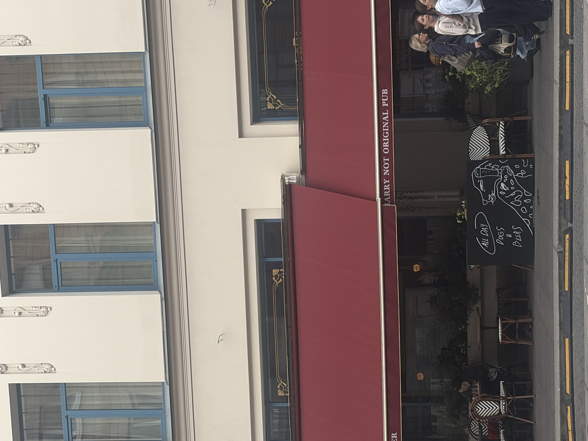 The image shows a storefront in Saint Petersburg, Russia. The building has a light-colored facade with ornate architectural details and blue-framed windows on the upper level. Below, a red awning extends over the storefront, which features large windows and outdoor seating. A sign above the awning reads "LARRY NOT ORIGINAL PUB". To the left, a person is seated at an outdoor table. To the right, a chalkboard displays the text "ALL DAY DOGS & BEERS" along with a drawing of two dogs. Several people are walking past the pub on the sidewalk, including two women and a girl. The street in front of the pub has yellow and gray traffic separators.
