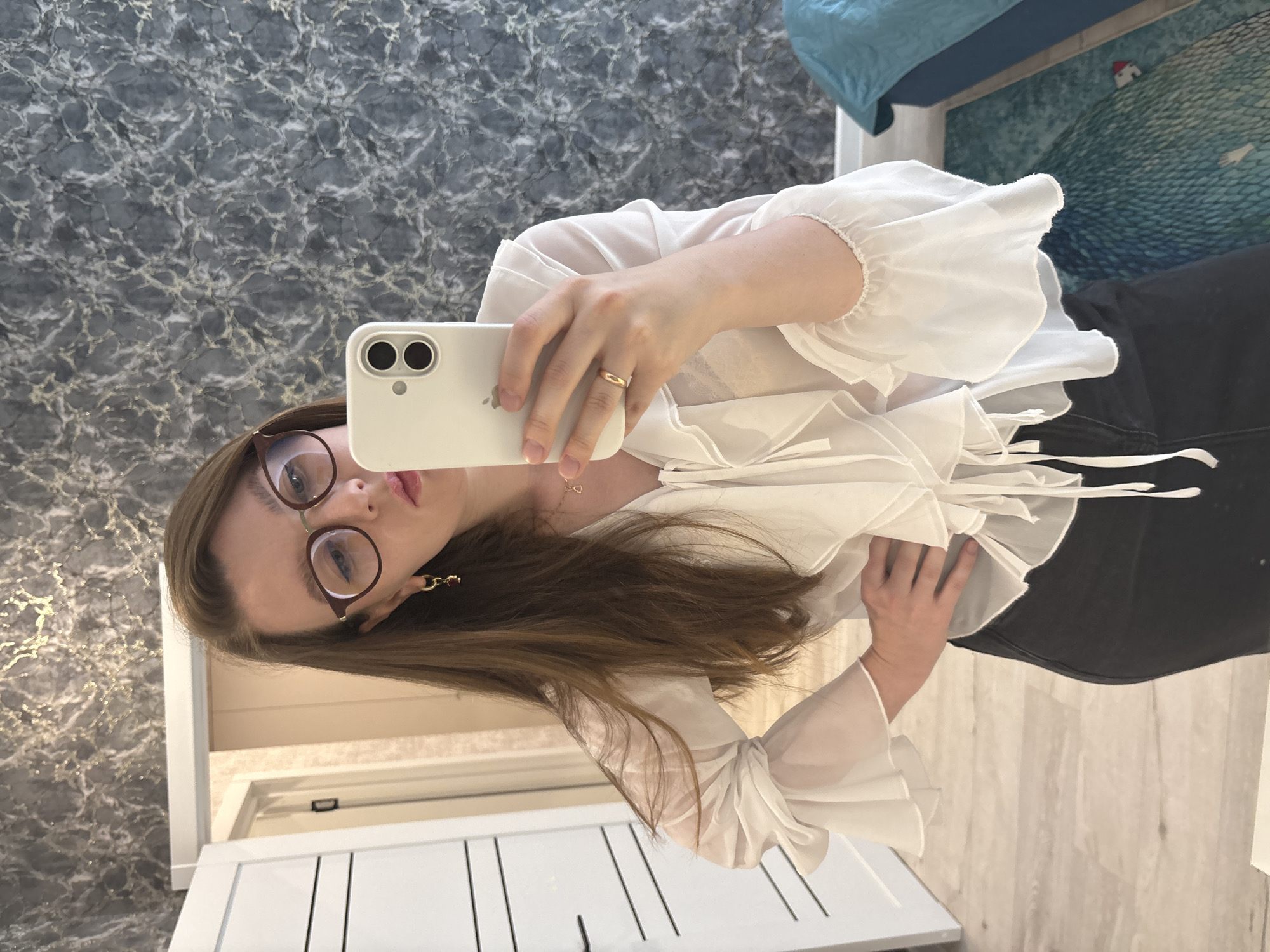 A person is taking a selfie in what appears to be an indoor setting. The person is wearing glasses and a white blouse. The setting appears to be a residential space with a marble-patterned wall, a wooden dresser, and a section of a blue bed visible in the background. There is no evidence to indicate this image was taken in the city of Izhevsk, Russia.