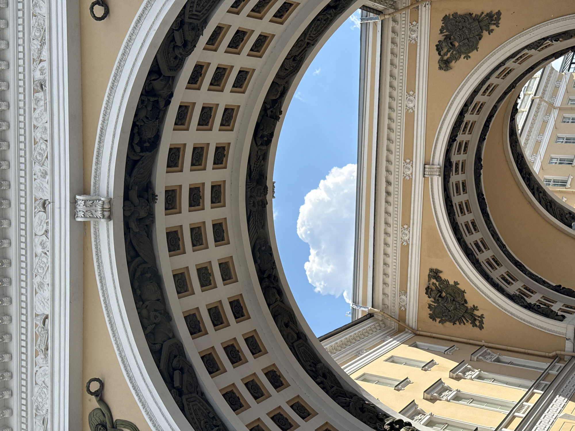 An upward-looking, tilted perspective shot captures the intricate classical architecture of a grand arched passageway, likely part of a historical building in Saint Petersburg, Russia. The structure features warm, pale yellow walls contrasted with ornate white decorative elements.
The foreground is dominated by two concentric, semicircular arches. The inner arch forms a coffered ceiling with numerous square panels, each containing a dark, rosette-like design. Along the inner curve of this arch are detailed dark relief carvings depicting classical figures or mythological scenes. The outer arch is also richly decorated with white moldings and continuous patterns.
Beyond the arches, a bright blue sky with a large, fluffy white cloud is visible, indicating clear, fair weather and bright daylight, likely midday or early afternoon. On the yellow wall segments between and around the arches, additional white and dark decorative elements are present, including wreaths, floral motifs, and, notably on the right side, bronze-colored military trophies featuring shields and weaponry.
Through the archway on the right, parts of another yellow building with white window frames are visible, suggesting either an adjacent structure or a building across an open space like a courtyard or square. The overall impression is one of majestic scale and elaborate classical ornamentation. No people or visible text are present in the image.