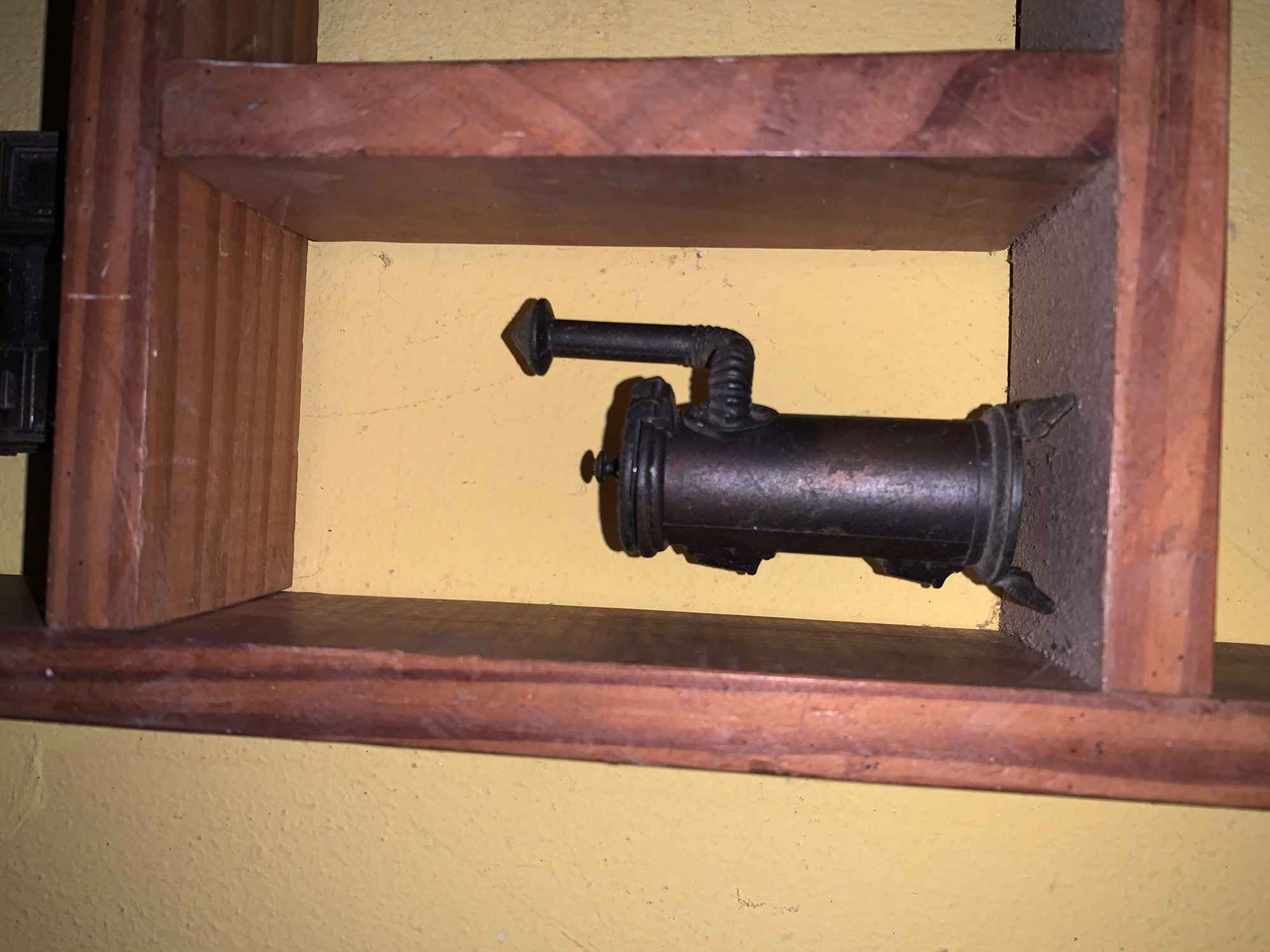 A cylindrical, metallic object, potentially an antique kerosene or gas lamp, rests within a simple, wooden shelf unit. The shelf is constructed of horizontal planks joined to vertical supports. The setting appears to be indoors against a yellow wall. The location is unknown, as Santa Ana, Venezuela is not evidenced. No people or interactions are depicted.