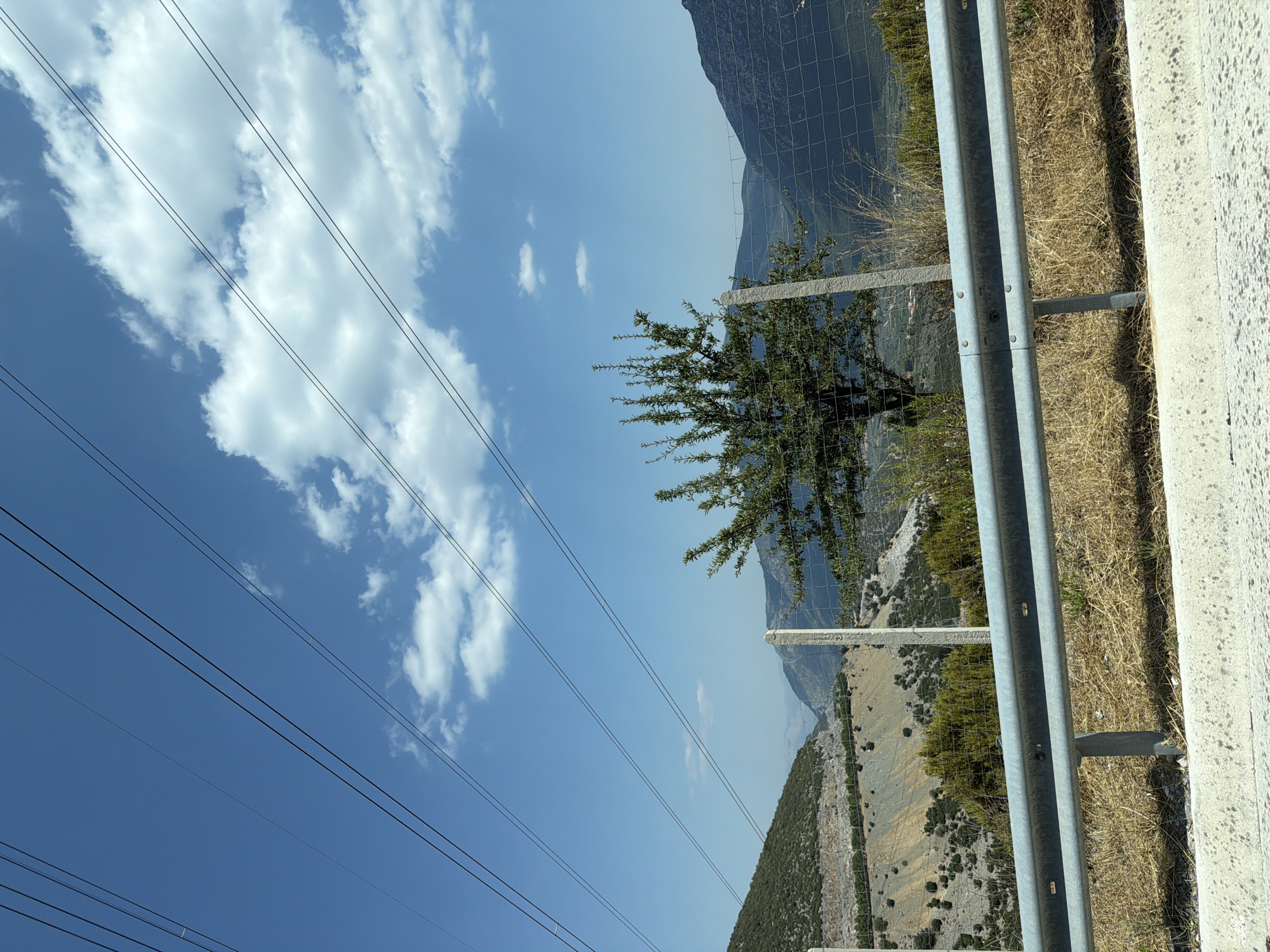 The image depicts a roadside view of a mountainous landscape under a clear blue sky with scattered white clouds. Power lines are prominently visible stretching across the upper portion of the frame. In the foreground, a metal guardrail runs horizontally along the bottom of the image, with dry grass and vegetation visible behind it. A wire fence with concrete posts is situated in the midground, separating the roadside from the natural environment. A small, leafy tree stands behind the fence, its branches reaching upwards. The background reveals a rugged, layered mountainous terrain, with evidence of a quarry or open-pit mine on the left slope. The overall impression is one of a rural, natural setting in Greece, possibly near the Municipal Unit of Lyrkeia.