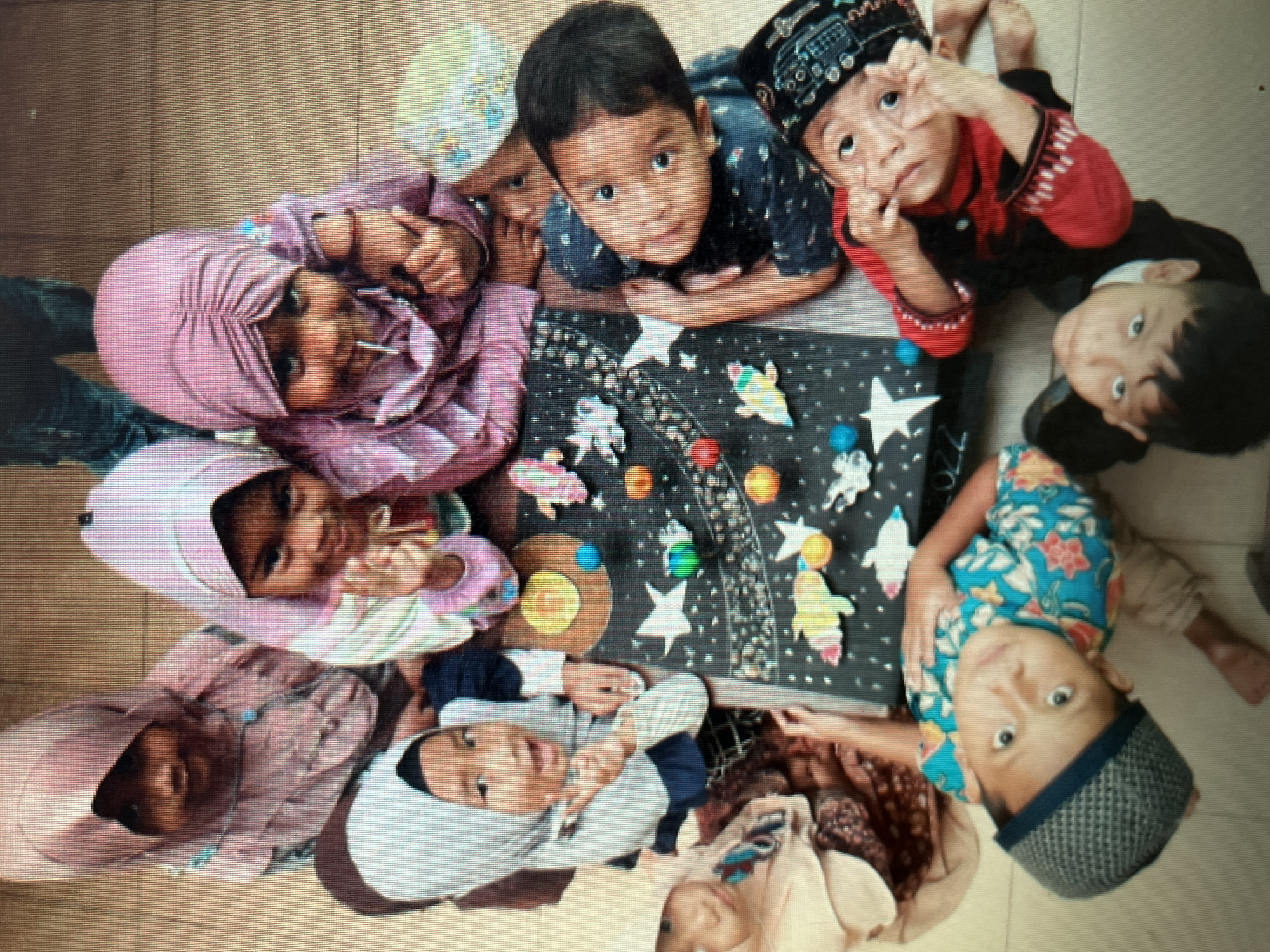 An overhead, slightly angled, medium shot captures a group of nine young children, likely preschoolers, gathered around a table in what appears to be a classroom setting. The children are of varying ethnicities, all wearing traditional Indonesian clothing, with several girls sporting hijabs. They are engaged with a black poster board adorned with colorful cutouts of stars, planets, and rockets, suggesting an educational activity related to space. Some children are pointing at the display, while others are interacting with each other. The floor is tiled, and a blurred background suggests the presence of other people and objects, possibly part of the school's common area. The lighting is bright, likely from overhead classroom lights or natural sunlight. The overall atmosphere appears to be one of joyful learning and engagement. The image is likely from a school in the city of Sejahtera, Indonesia, given the context of the children's attire and the implied location.