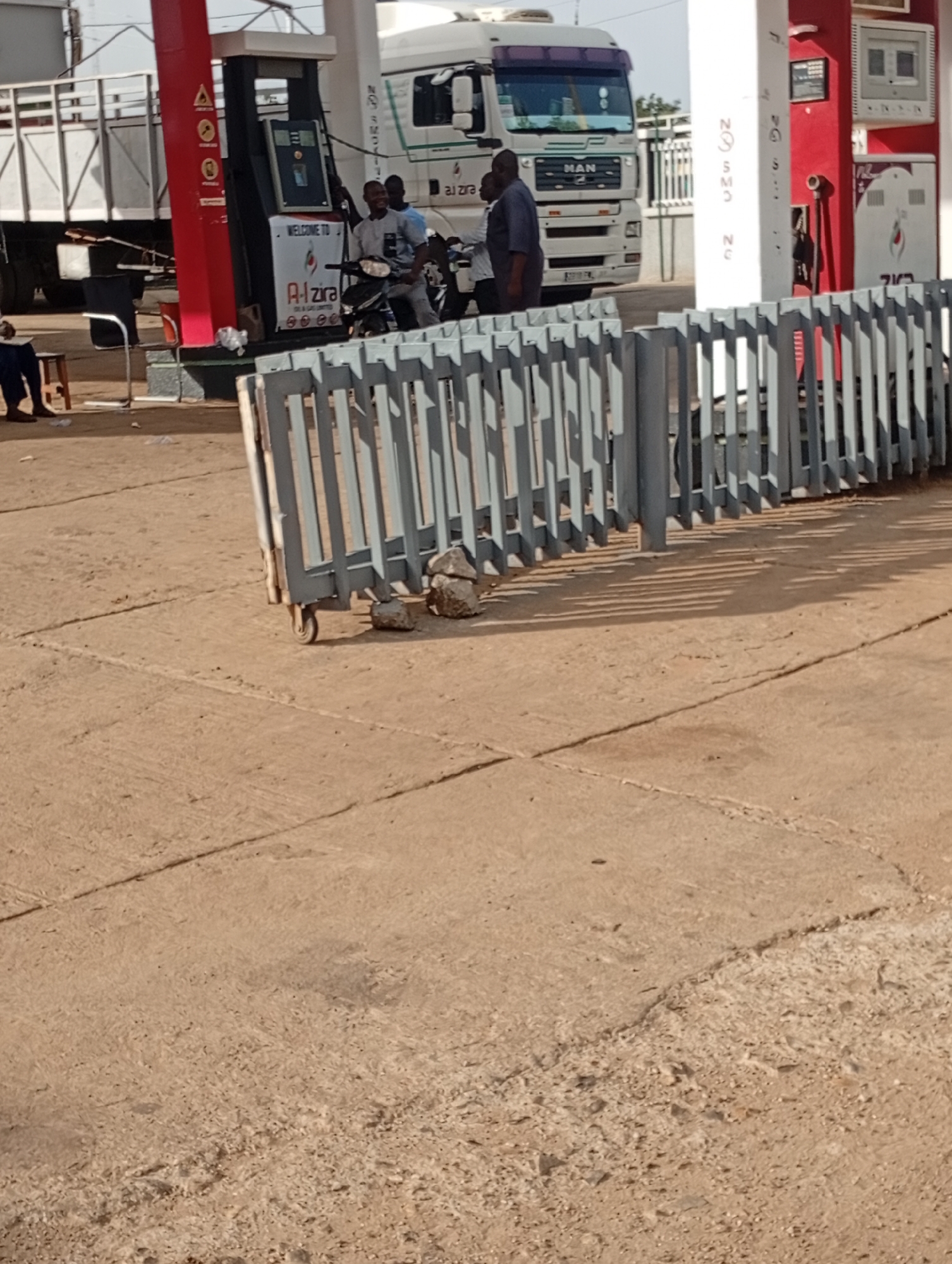 At a gas station in Rafin Par, Nigeria, several people are present. A white semi-truck with "MAN" written on the front is parked near a gas pump. Three individuals are gathered around a motorcycle, one of whom is operating it. Another person is seated on a stool to the left. A grey metal gate is positioned in the foreground, with a wheel visible on its right side. The ground is paved with concrete, showing cracks and some debris.