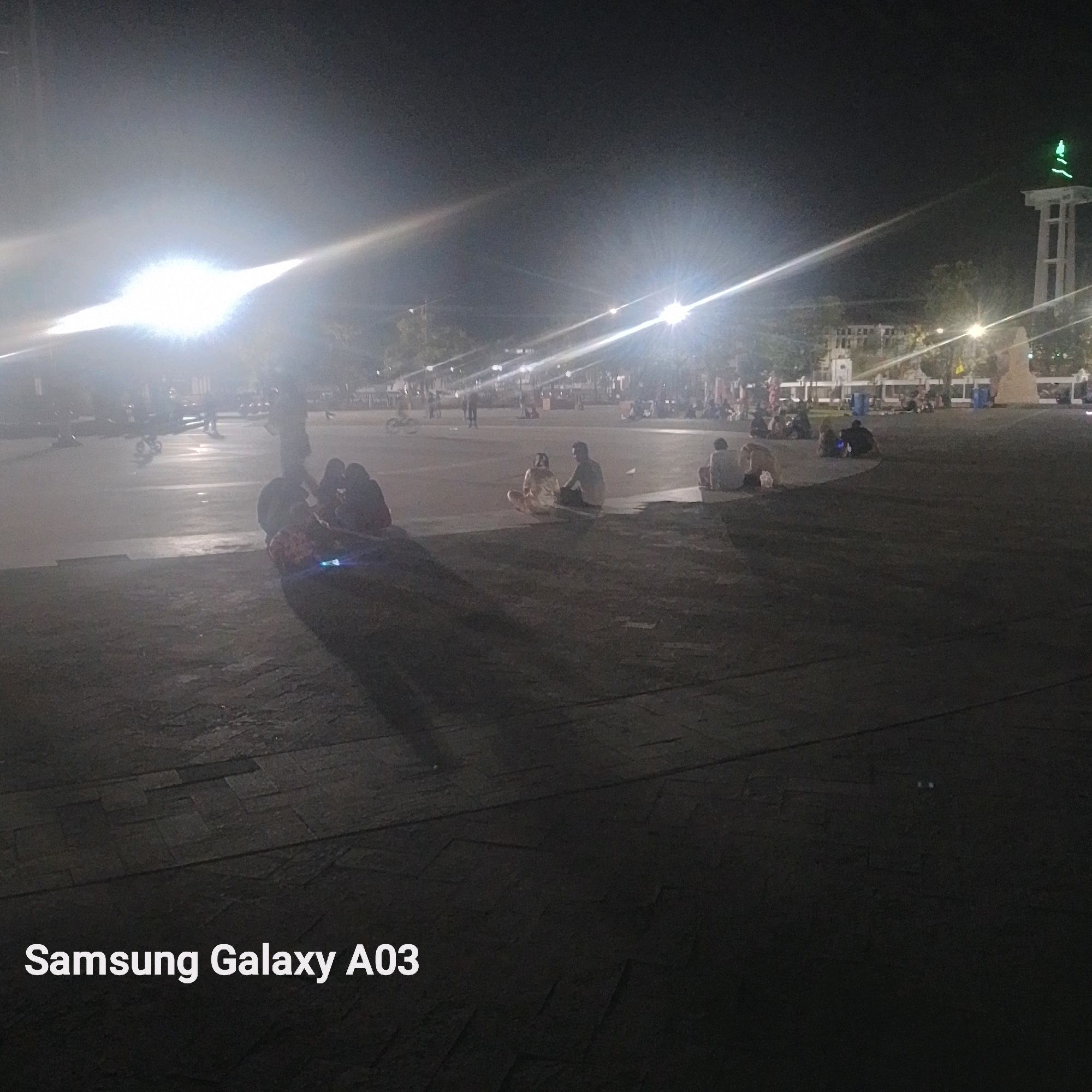 This media file captures a nighttime scene in what appears to be a public square or park in Indramayu, Indonesia. The image is dominated by artificial lighting, with bright spotlights creating lens flares and casting long shadows across the textured ground.
Several people are visible, mostly sitting or lying down on the ground, appearing relaxed and engaged in conversation or simply enjoying the evening. Some groups are huddled together, while others are more spread out. The activity seems to be a casual gathering of people in a public space at night.
Notable details include a tall, tower-like structure in the background with a green light at its peak, and other illuminated structures that suggest a built environment. The time of day is clearly nighttime due to the darkness and artificial lighting. The overall mood appears peaceful and social, with people spending their evening outdoors.
The text "Samsung Galaxy A03" is visible in the bottom left corner of the image, indicating it is a photograph taken with that particular device.