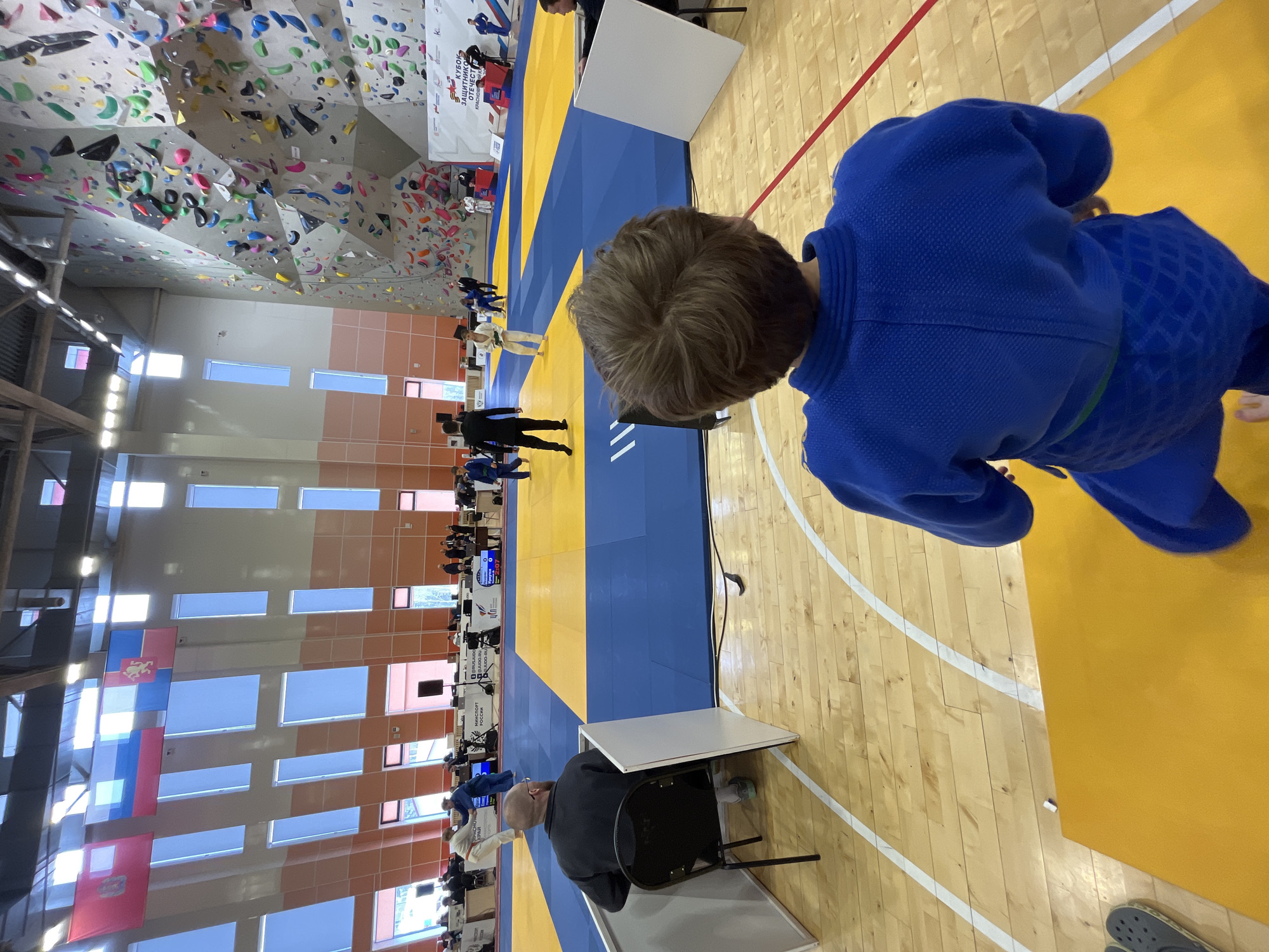A photo taken indoors in the city of Krasnoyarsk, Russia, at an athletic event. A boy in a blue gi stands on a wooden floor next to a blue and yellow mat. On the mat are multiple people, primarily wearing gis, participating in a judo tournament. In the background is a climbing wall. The walls have orange and white sections, and multiple tall windows. There are Russian flags hanging. A man sits at a table on the right, facing the tournament.