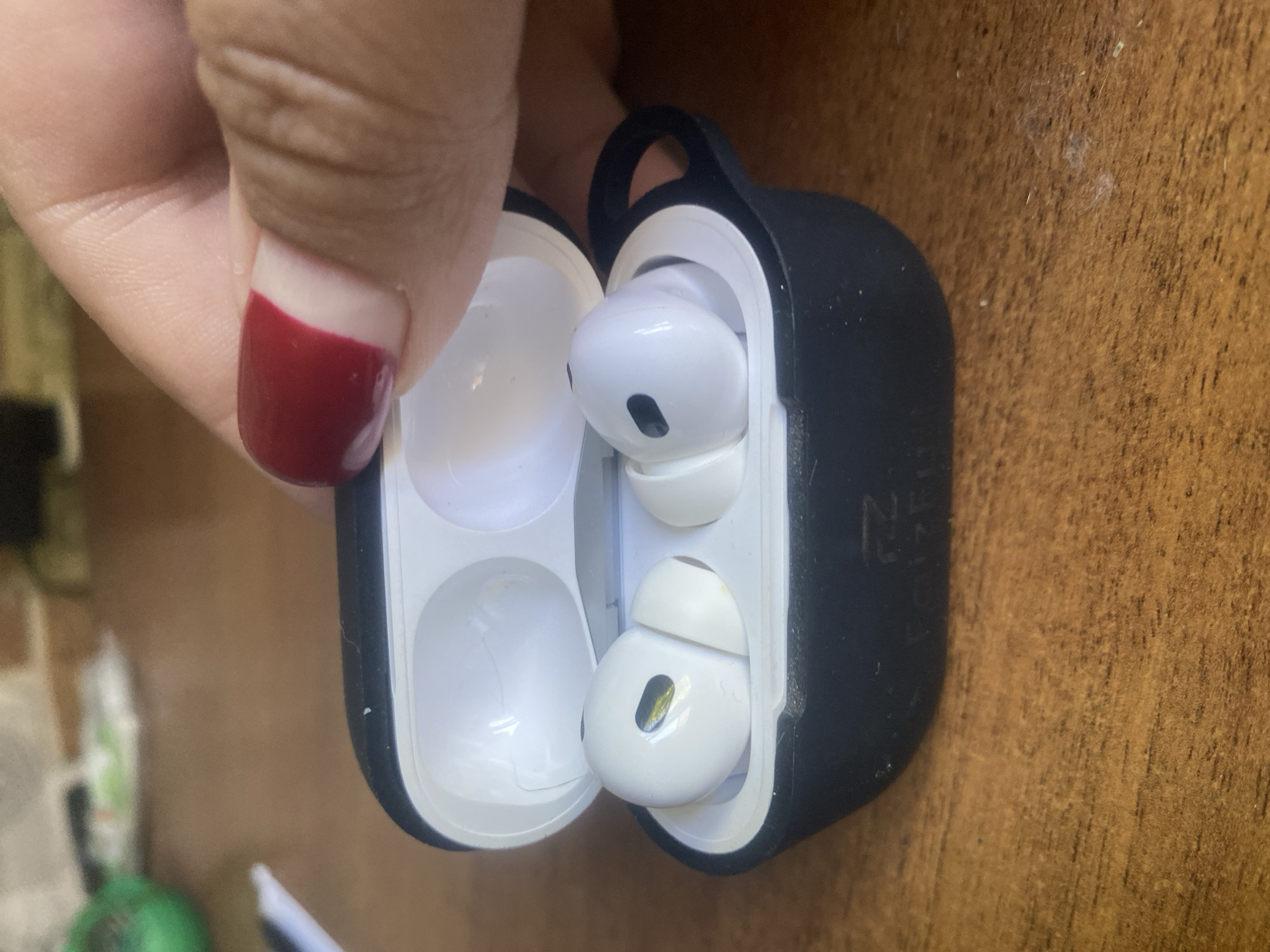 A close-up shot shows a pair of white wireless earbuds in a black charging case. The case is open, revealing the earbuds nestled inside. A person's hand with a fingernail painted dark red is visible, gently holding the top of the case. The earbuds have black mesh grilles. The background is a wooden surface, and slightly out of focus in the upper left corner, there appear to be some green and white objects. The image appears to be an indoor scene, with no specific indications of the city of Saint Petersburg, Russia.