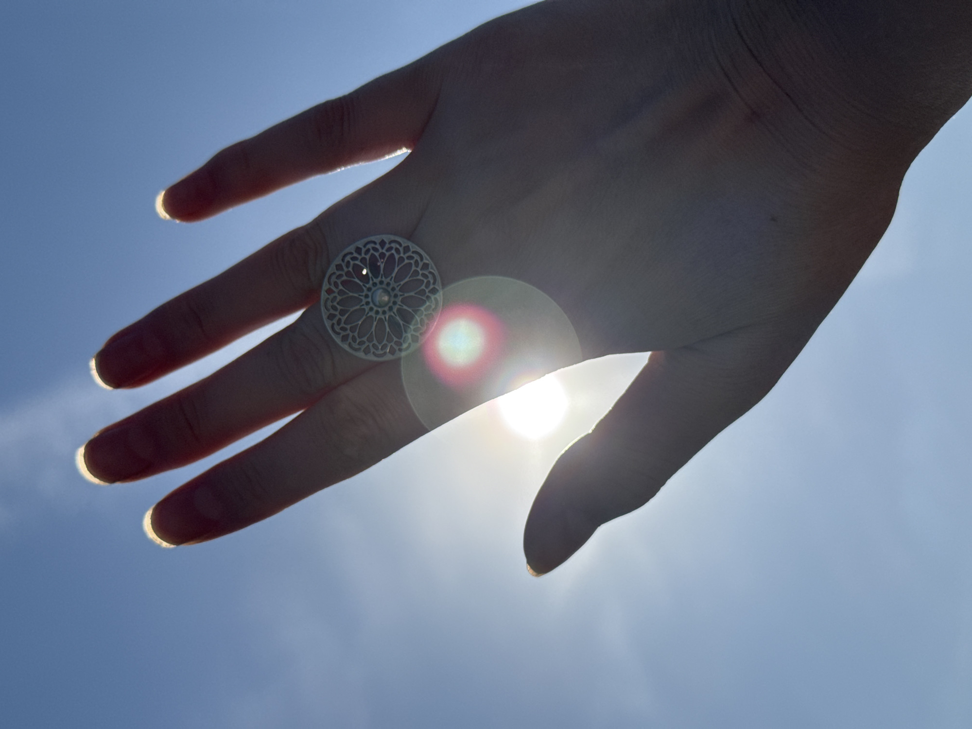 A hand, viewed from below against a bright sky, dominates the frame. The hand is positioned in front of the sun, creating a lens flare effect. A large, ornate, circular ring is on the middle finger. The background is a clear, light blue sky. The location is not identifiable as Izhevsk, Russia based on the image content alone.