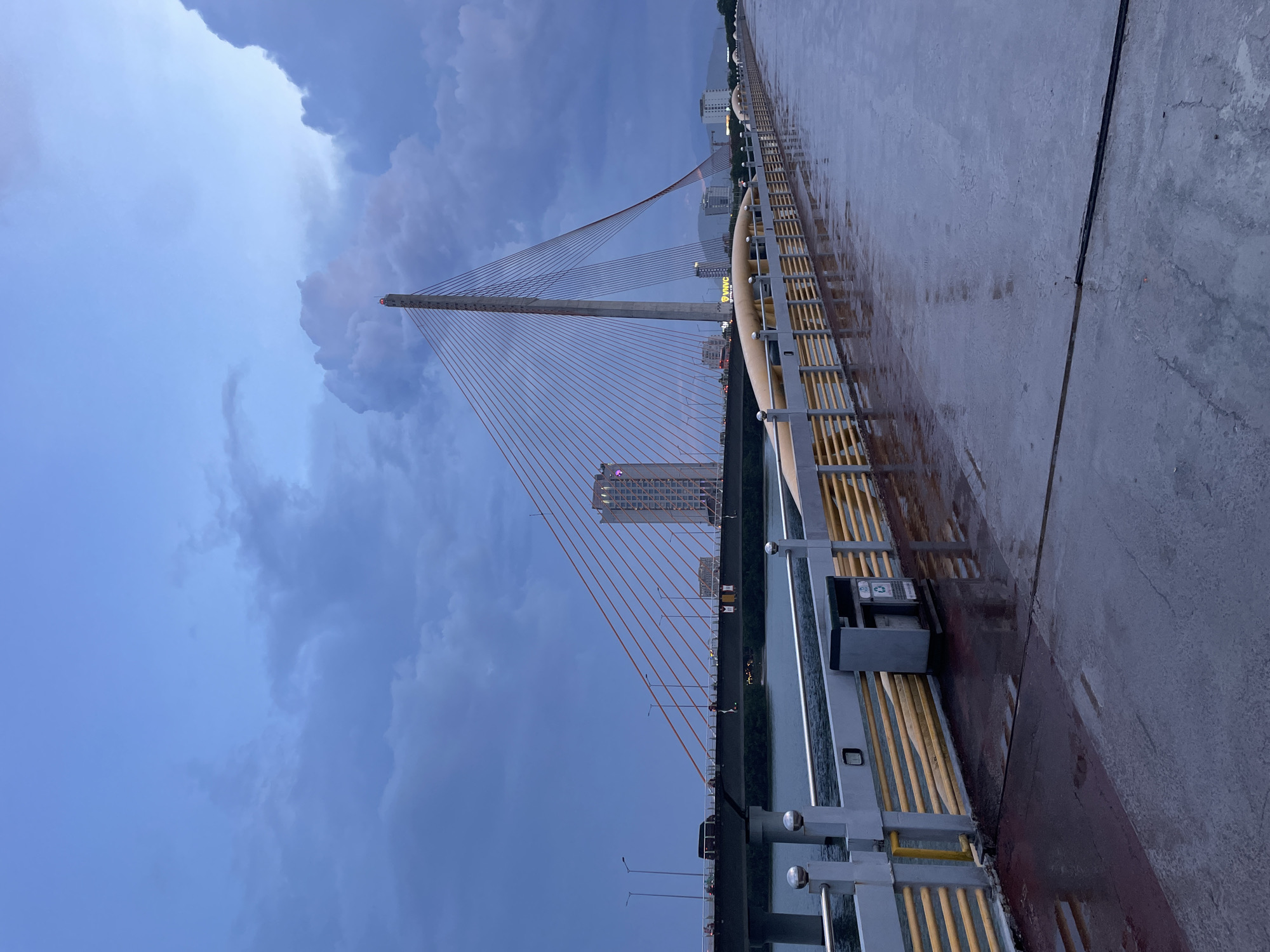 This is a vertical, eye-level shot of a modern cable-stayed bridge at dusk, likely in Da Nang, Vietnam, given the location context. The sky is a dramatic mix of deep blues and grays, with large, brooding storm clouds dominating the upper left portion of the frame. The bridge's concrete pylon rises majestically, adorned with numerous thin, orange cables that fan out to support the bridge deck.
The bridge deck itself is wet and reflective, suggesting recent rain or the aftermath of a storm. A yellow and gray railing system runs along the side of the pedestrian walkway, which is the vantage point for the photograph. Beside the railing, a dark gray trash receptacle with a recycling symbol is visible. The walkway appears to be made of reddish-brown asphalt, also wet and shimmering.
In the background, across the body of water the bridge spans, a cityscape is visible. Buildings of varying heights line the riverbank, with the most prominent being a tall, modern structure with visible windows, possibly an apartment or office building. The lights of the city are beginning to twinkle as daylight fades. The overall mood is somber and dramatic, amplified by the stormy sky and the wet conditions. The image captures a sense of anticipation, perhaps for the storm to fully arrive or to pass. No people are visible in the frame.