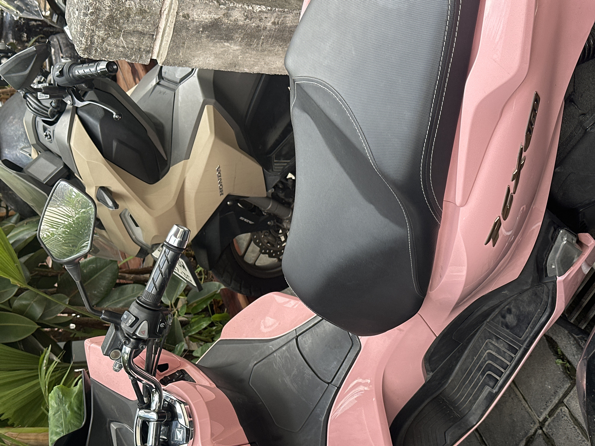 Two Honda scooters are parked in North Kuta, Indonesia. In the foreground, a pink scooter is parked, showing its front handlebars, seat, and leg rest. A beige scooter is parked behind it. The scooters are parked on a paved surface, with a concrete pillar and lush green foliage visible in the background.