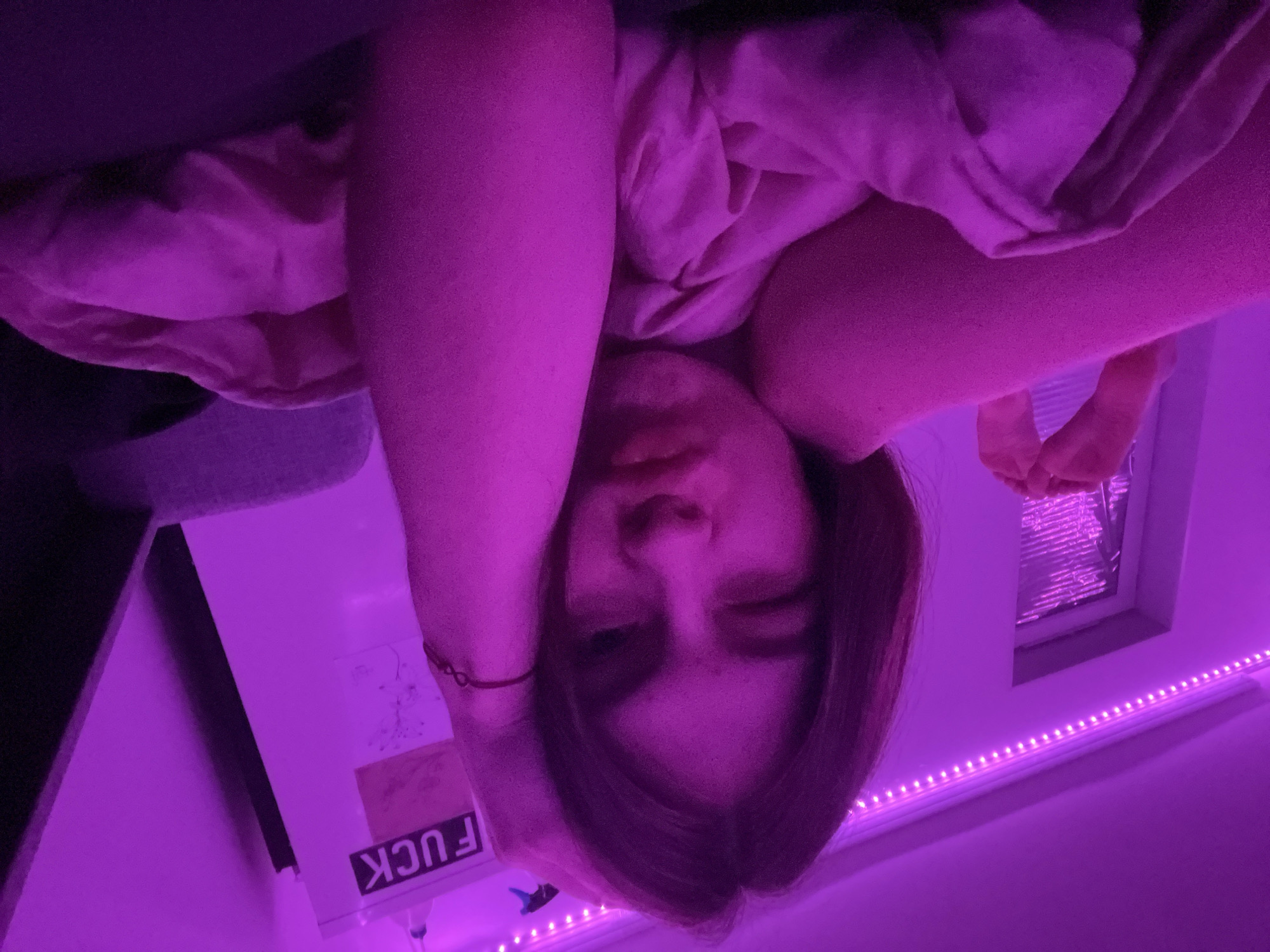 The image presents a unique, high-angle portrait of a young person lying down, with their head appearing inverted due to the camera's perspective. The subject's face is central, with partially closed eyes and slightly parted lips, conveying a relaxed or contemplative mood. Their arms are raised, resting around their head, while their shoulder-length, dark hair frames their face. A red string bracelet is visible on their left wrist.
The entire scene is bathed in a vivid purple and pink glow, originating from LED strip lights positioned along what appears to be a wall or ceiling edge in the background. This artificial illumination suggests the photo was taken during the evening or night, within an intimate indoor setting, likely a bedroom in Saint Petersburg, Russia.
In the upper right portion of the frame, a window or opening is partially visible, appearing to be covered by blinds or a reflective material, with a pair of bare feet or lower legs positioned near it. To the left, a surface or shelf is visible, featuring various items and a white object with the word "FUCK" printed prominently and upside down. The person's body is partially obscured by light-colored, rumpled clothing or blankets, blending with the dominant purple hues. The unconventional angle creates a personal and artistic self-portrait.