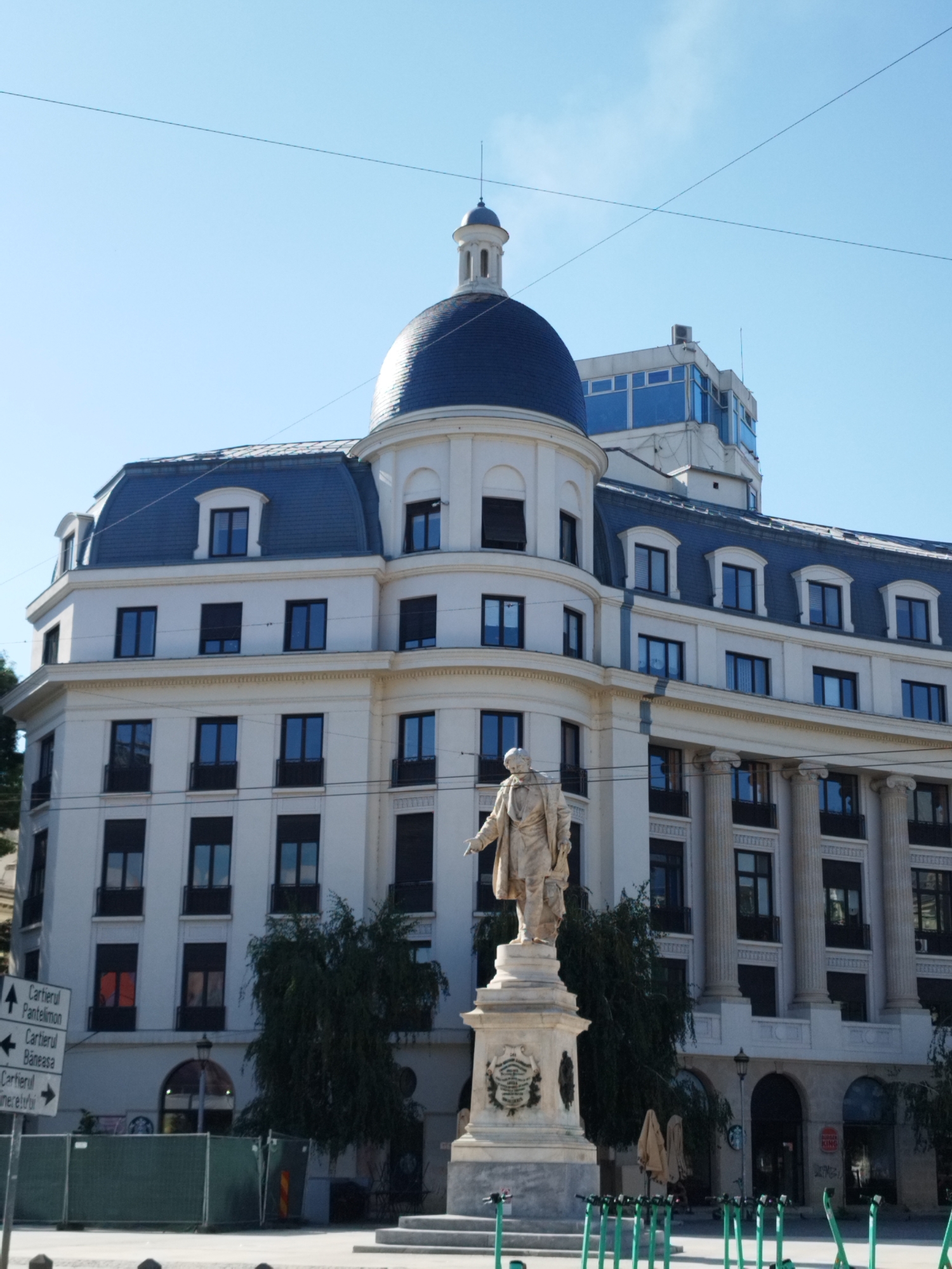 This image captures a frontal view of the National Bank of Romania building in Bucharest, Romania, on a clear, sunny day. The architectural centerpiece is a multi-story white building with a prominent dark blue domed roof on the left side, topped with a spire. The building features numerous dark-framed windows and a classical facade with columns on the right. In the foreground, a stone statue of a man in historical attire stands on a pedestal, with the base of the statue featuring an inscription and a circular emblem. Several green electric scooters are parked in rows on the paved ground in front of the building. The background is dominated by a bright blue sky with thin, high-altitude lines of utility wires crisscrossing it. Signs indicating directions to various districts like "Cartierul Pantelimon," "Cartierul Baneasa," and "Cartierul Unirii" are visible on the left. The scene appears to be a public square or street in a downtown area.