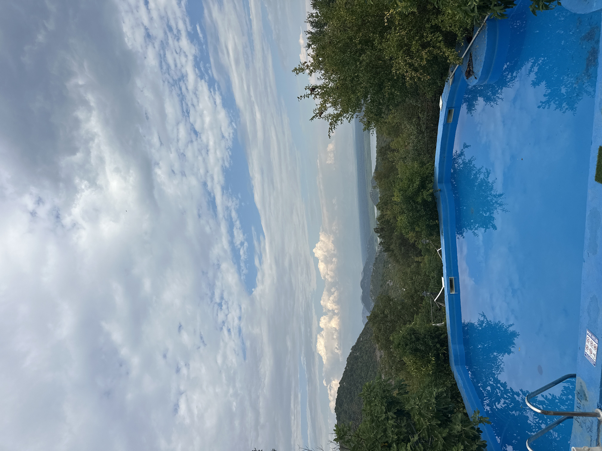The image depicts an outdoor swimming pool with vibrant blue water, reflecting the sky and clouds. The pool is situated on an elevated vantage point, offering a panoramic view of a landscape characterized by rolling hills covered in lush green trees. In the distance, a body of water, likely a lake or sea, is visible, along with more distant hills under a sky filled with scattered clouds. The foreground features a portion of the pool's edge with metal ladder rungs visible. The overall scene suggests a tranquil setting in a natural environment. This scene is located in the city of Cetinje, Montenegro, known for its mountainous terrain and proximity to Skadar Lake.