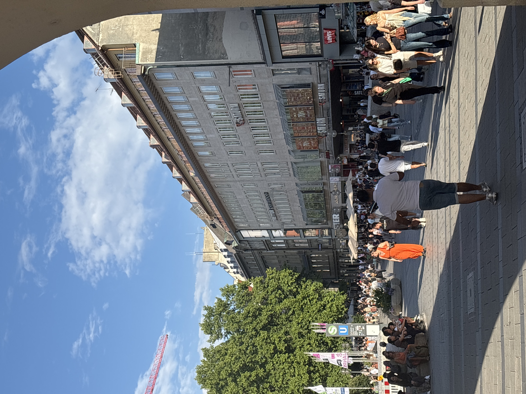 The image depicts a bustling pedestrian street in Munich, Germany, likely in the city center, under a clear blue sky with scattered clouds. The scene is dominated by a large, multi-story building with a grey facade, featuring numerous windows and signs for businesses, including "SATURN" and "Levi's." A construction crane is visible in the upper left corner, suggesting ongoing development in the area.
The street is paved with stone tiles and is filled with people walking, shopping, and socializing. Many individuals are dressed in casual summer attire. Some people are seated on benches along the side of the street, while others are browsing shops. Several outdoor cafes with umbrellas are set up, indicating a lively commercial and social atmosphere.
In the foreground, a man in a white t-shirt and blue shorts walks away from the camera, casting a long shadow on the pavement. To his left, a group of people are seated on benches. Further down the street, a woman in a bright orange dress stands out against the crowd. The overall impression is one of a vibrant urban environment on a sunny day. The presence of the "U" symbol indicates a U-Bahn (subway) station, likely Karlsplatz (Stachus) given the context of a busy shopping street in Munich.