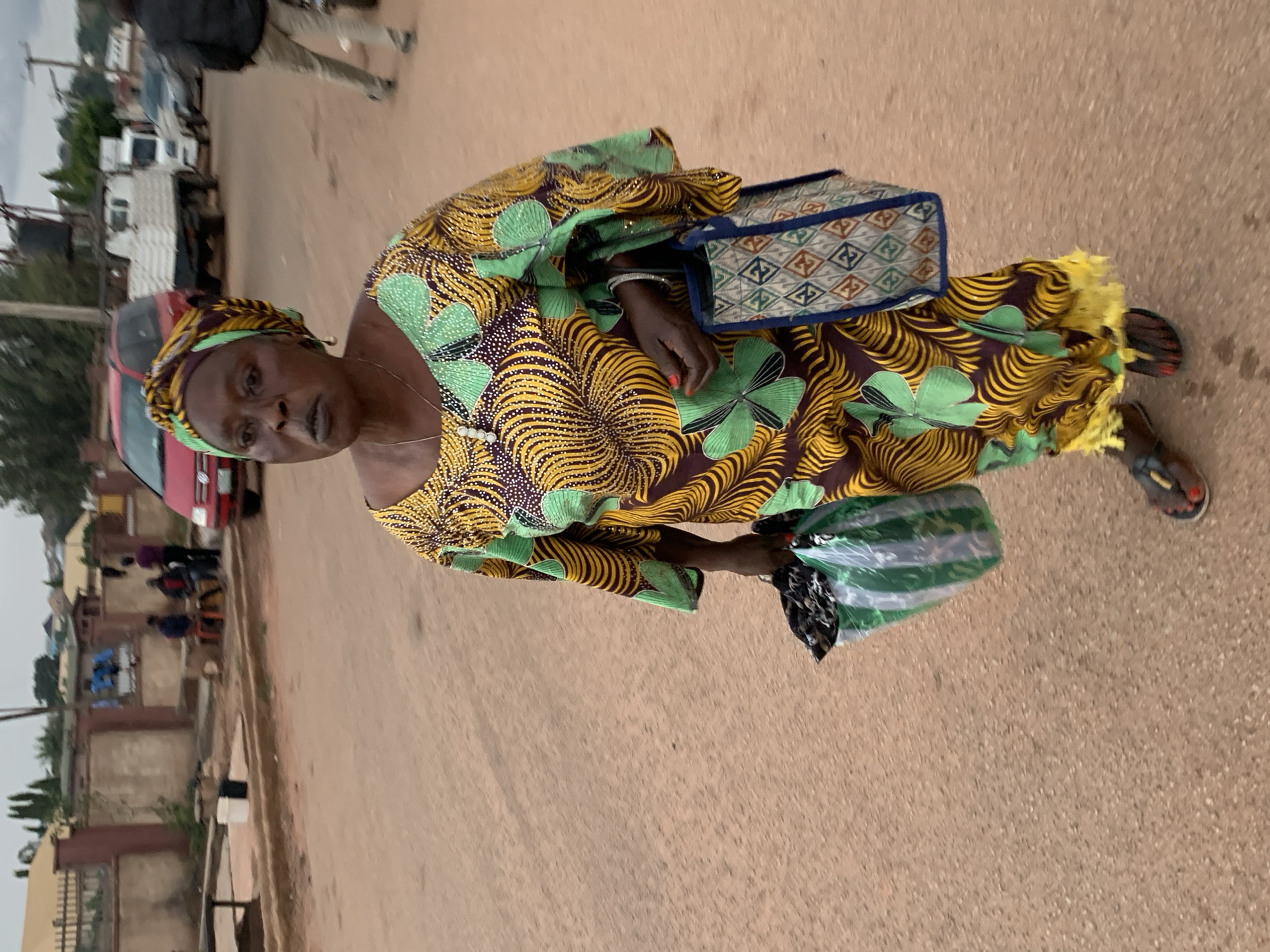 A woman with dark skin, wearing a colorful yellow, green, and brown patterned dress and headscarf, walks on a dirt road in Jos, Nigeria. She is carrying two bags, one a green and white plastic bag in her left hand and a patterned tote bag in her right. A few people are visible in the background, along with parked vehicles, including a red car and a white truck. The environment appears to be a typical street scene in a Nigerian city.