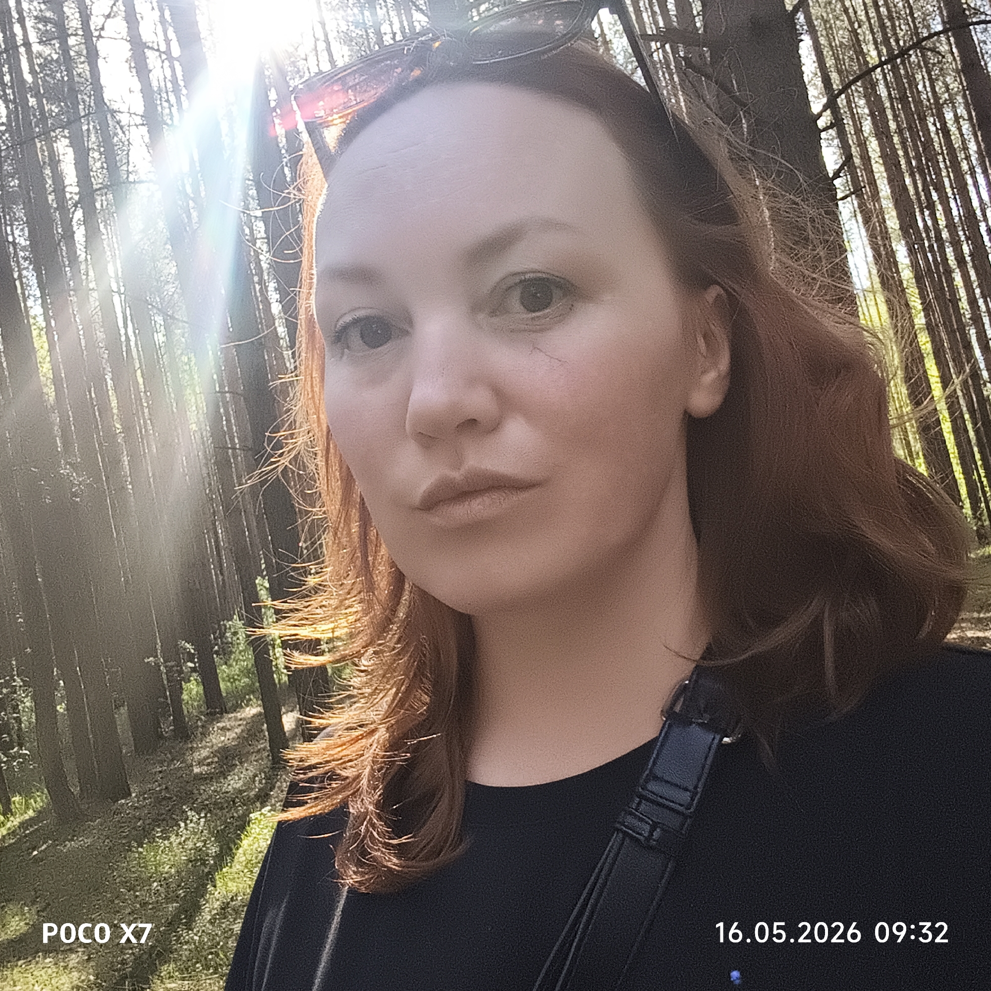 A selfie was taken in a wooded area near Chaikovsky, Russia. A person with reddish-brown hair is in the foreground, wearing sunglasses on their head and a black shirt with a black bag strap across their shoulder. The background consists of tall, thin trees with sunlight filtering through the branches. The date and time "16.05.2026 09:32" is imprinted in the lower right corner, and the camera model "POCO X7" is imprinted in the lower left corner.