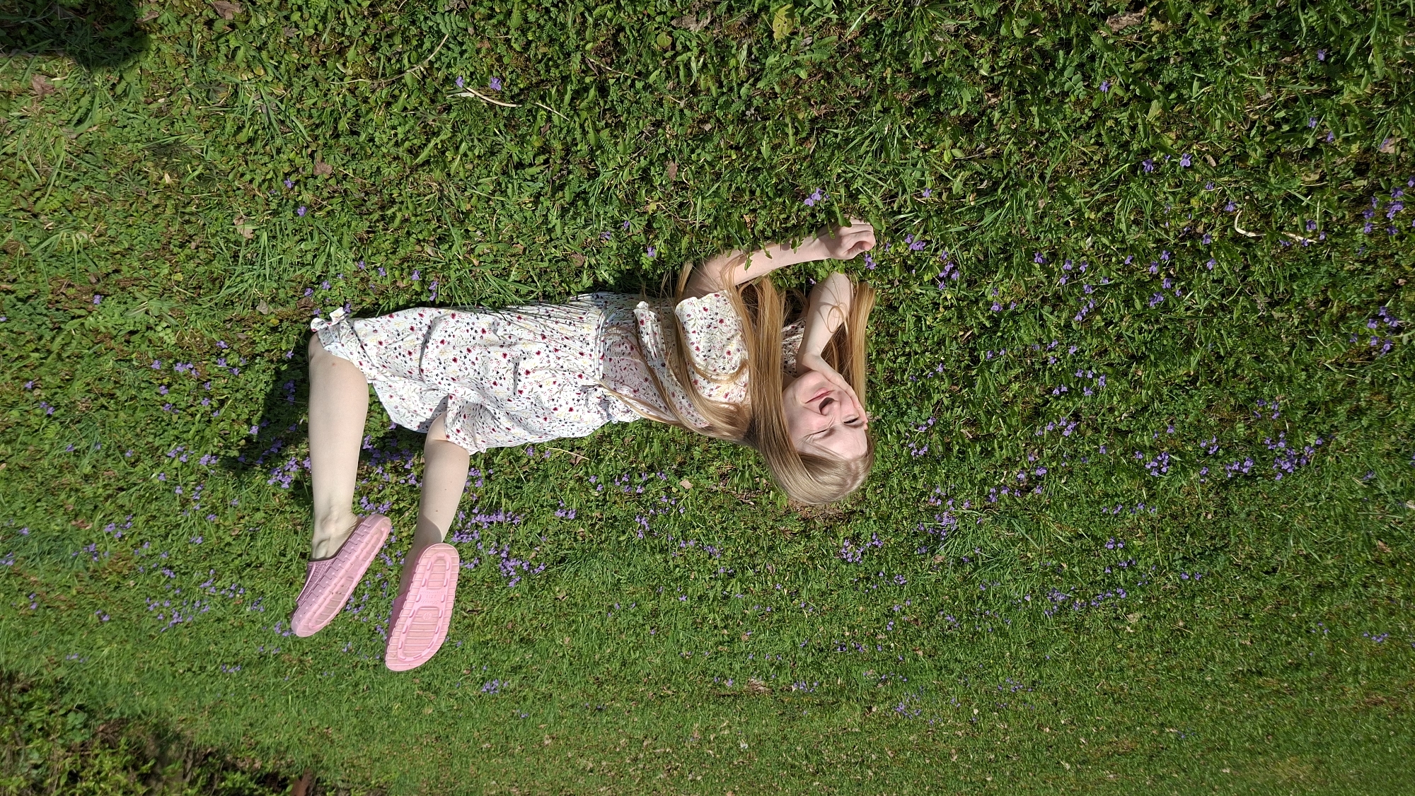 A young woman is lying on her back in a grassy field in Большой Букор, Russia. She is wearing a light-colored floral dress and pink sandals. Her blonde hair is spread out on the grass. Small purple flowers are scattered throughout the field. She is smiling, with her arm bent behind her head.