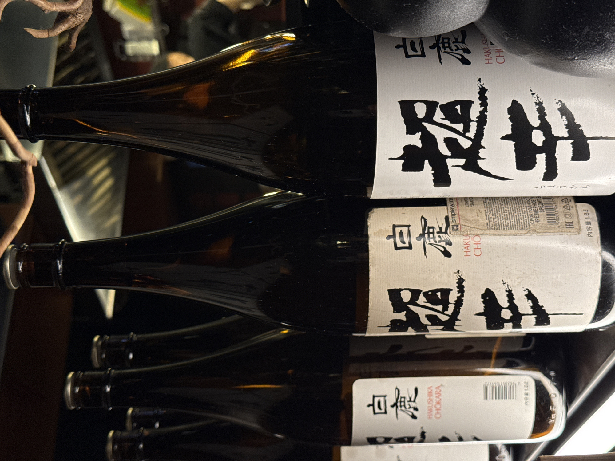The image displays a close-up view of several bottles of sake, stacked vertically. The bottles are made of dark glass and have white labels with Japanese calligraphy and some text in English and Russian. The Japanese characters appear to be "白鹿" (Shiraka), which translates to "White Deer," a brand of sake. Below the calligraphy, the text "HAKUSHIKA CHOKARA" is visible, indicating the type or name of the sake.
In the background, there are out-of-focus elements that suggest an indoor setting, possibly a bar, restaurant, or a retail display. The lighting is somewhat dim, with highlights reflecting off the glass bottles, suggesting artificial lighting.
One of the labels also contains Cyrillic text, "СРОК ГОДНОСТИ ЧРЕЗМЕРНОЕ У ВРЕДИТ В," which translates to "SHELF LIFE EXCESSIVE HARM TO." Another label shows "内容量 1.8ℓ," indicating a volume of 1.8 liters. A barcode is also visible on one of the labels. The overall scene is focused on the products themselves, with minimal emphasis on the surrounding environment. The context provided indicates the location is Moscow, Russia, which aligns with the presence of Russian text on the labels.