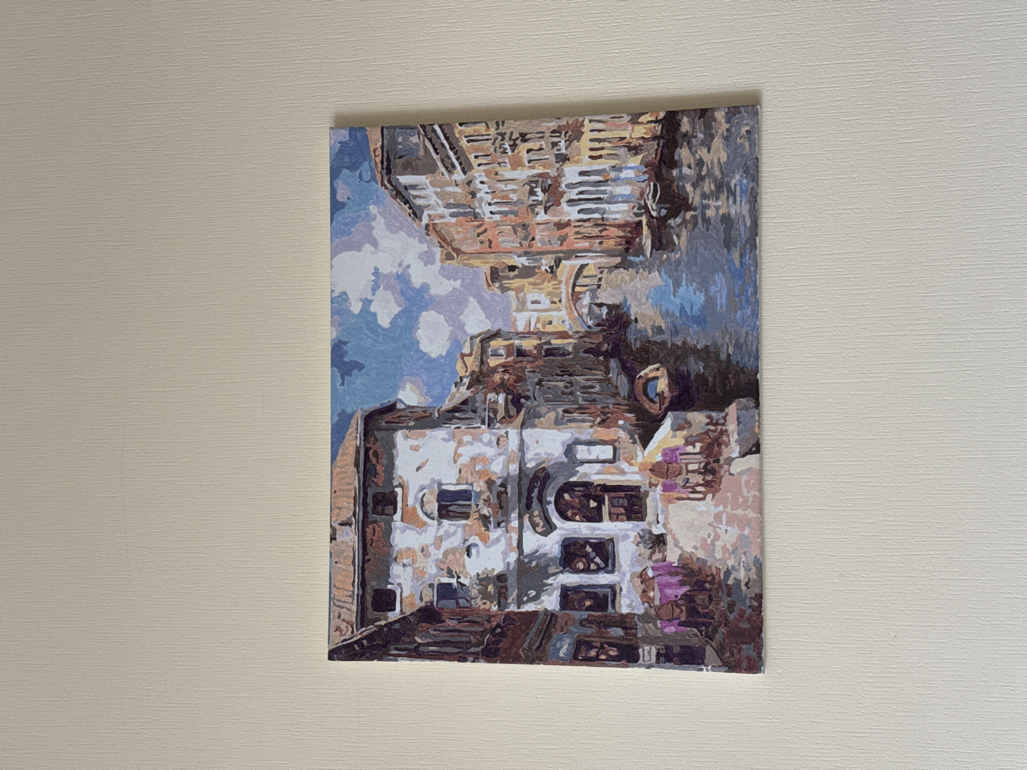 The media file displays a paint-by-numbers artwork, depicting an urban canal scene, hung vertically against a light beige, subtly textured wall. The image itself is rotated 90 degrees clockwise; when viewed correctly, the painting presents a vibrant interpretation of a cityscape, likely Saint Petersburg, Russia, given the provided context.
The artwork features a canal flanked by multi-story buildings under a partially cloudy sky. The painting employs a distinct, blocky style with areas of flat, unblended color, resembling a mosaic or pixel art. The sky, at the top of the painting, is rendered in shades of light and medium blue with abstract shapes of white and pale purple suggesting clouds.
The buildings lining the canal exhibit varied architectural details. One prominent building on the left (when correctly oriented) has a light-colored facade with visible signs of age or distress, featuring multiple dark-paned windows and some arched openings. Other buildings, particularly in the background and along the right side, show warmer tones of brown, orange, and beige, with intricate, blocky patterns suggesting bricks, balconies, or decorative elements. The water in the canal, depicted in dark blues and grays, reflects the surrounding structures in a similarly blocky, abstract manner.
The overall impression is of a daytime scene, though the lighting is soft and diffuse, without harsh shadows. There are no people visible, and no specific activities are taking place within the static scene. The painting itself is the primary object, showcasing a completed creative project. No text is visible in the artwork or on the wall.