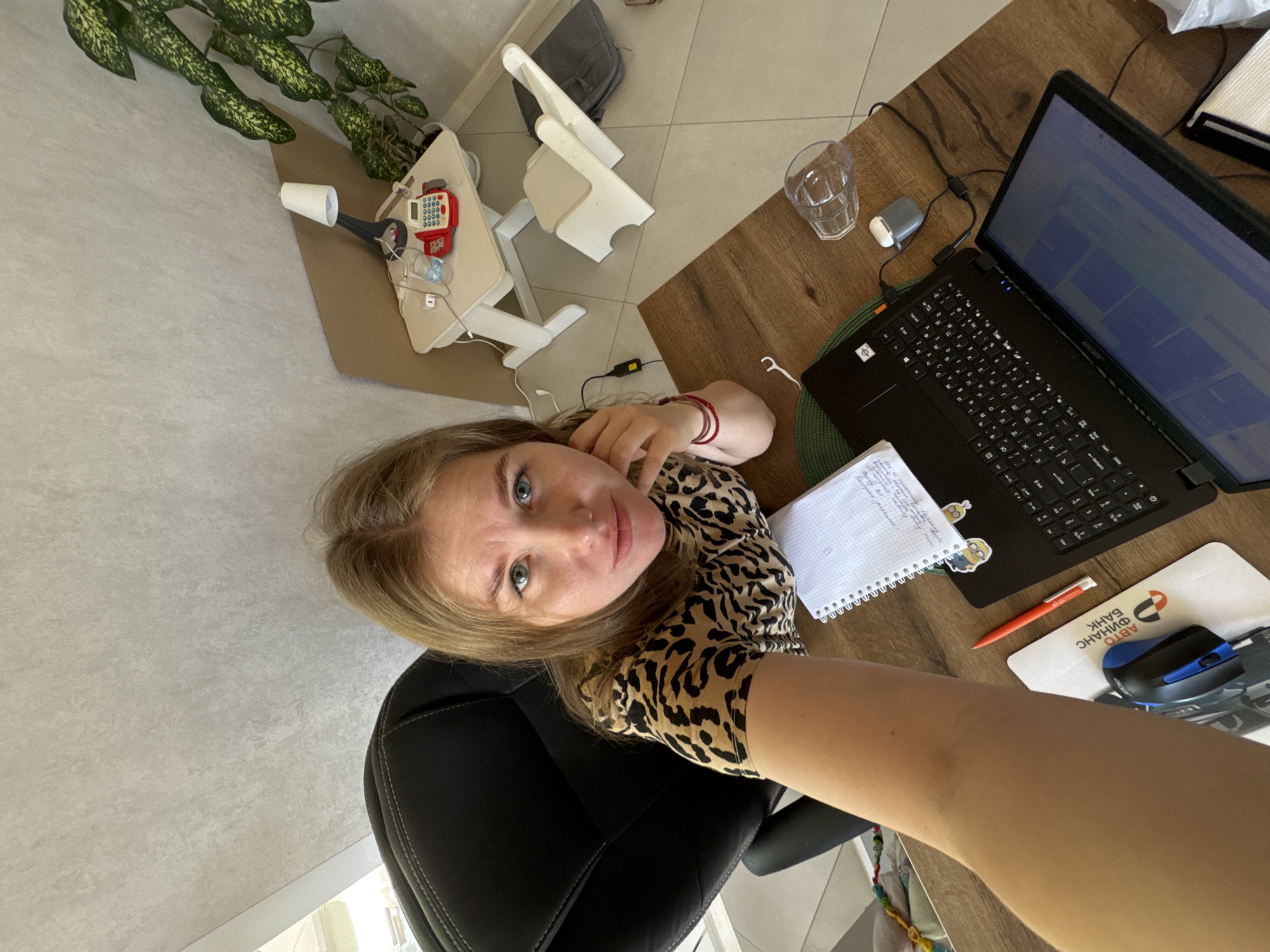 A woman is sitting in a black office chair, holding her arm out to take a selfie or video. She is fair-skinned with blonde hair and wearing a leopard-print top. She is positioned near a wooden desk.
On the desk, there is a black laptop, a notepad, a red pen, and a mousepad with a blue mouse. A glass of water and a pair of white wireless earbuds are also on the desk.
In the background, there is a tall green plant, a children's high chair, and a small white table with what appears to be a toy cash register and a white cup.
The location is Ферма, Russia.