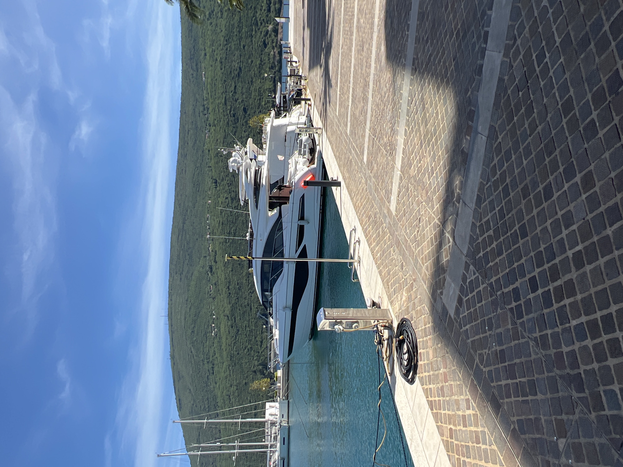 The image shows a marina in Herceg Novi, Montenegro, with a large white yacht docked alongside a stone quay. The yacht is sleek and modern, with multiple decks and a prominent radar array. The quay is made of dark cobblestones, with a lighter stone border along the water's edge. A black hose reel and a service pedestal are visible on the quay, indicating the availability of utilities for the boats. In the background, a steep, forested hillside rises towards a clear blue sky. Several sailboats with tall masts are also visible in the marina. The angle of the sun suggests it is either morning or afternoon.