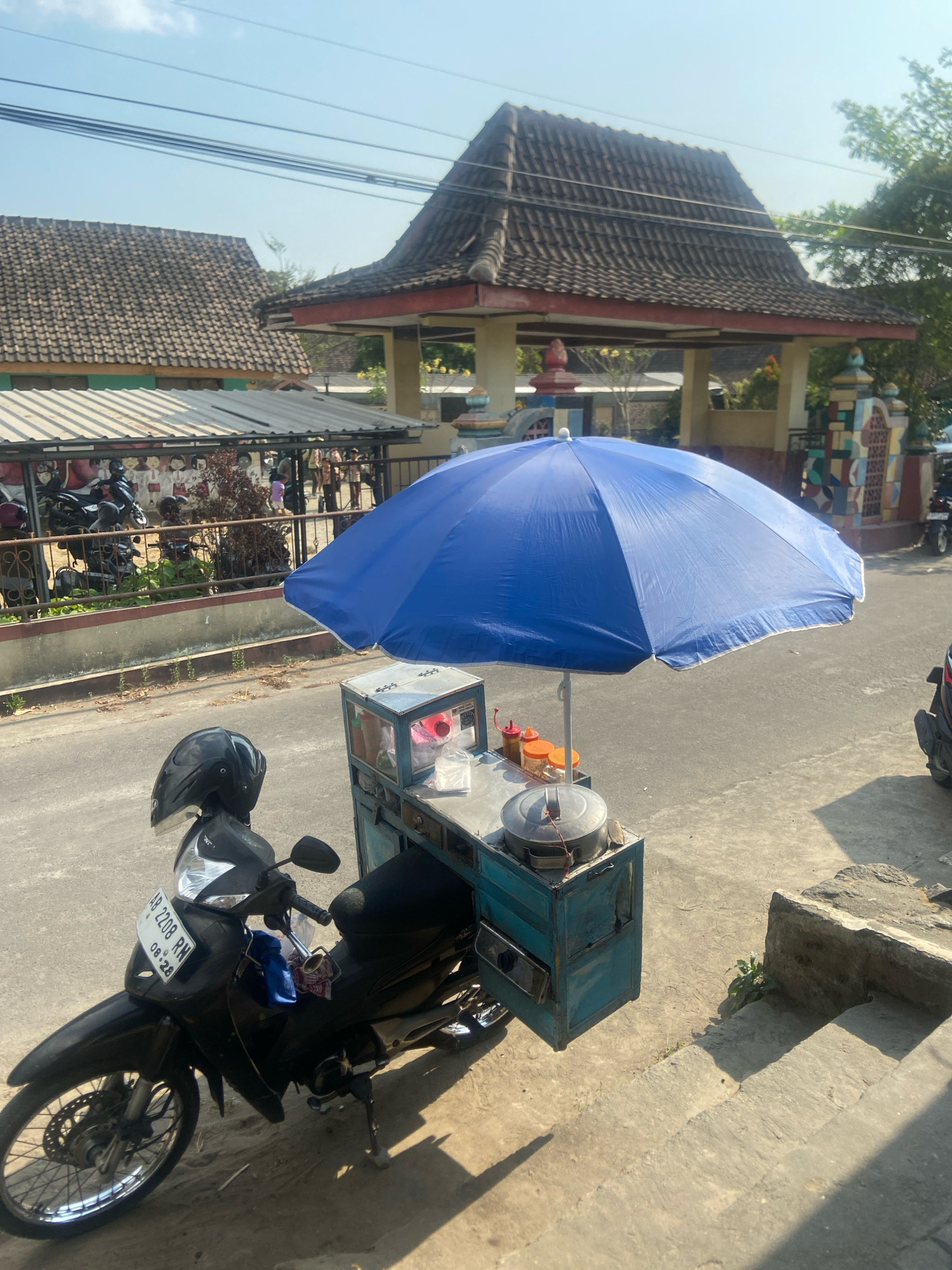 The image captures a street scene in Sleman, Indonesia, on a bright, sunny day. A black motorcycle with a license plate "AB 2208 RN" is parked on the side of the road, facing left. On the motorcycle's rear, a blue cart with a metal frame and a blue umbrella is attached. The cart appears to be a food stall, containing containers and condiments. In the background, several buildings with traditional tiled roofs are visible, along with trees and power lines. The street is paved, and a concrete sidewalk runs along the right side of the image. The overall impression is one of a typical day in a bustling Indonesian town, with street vendors and local architecture.