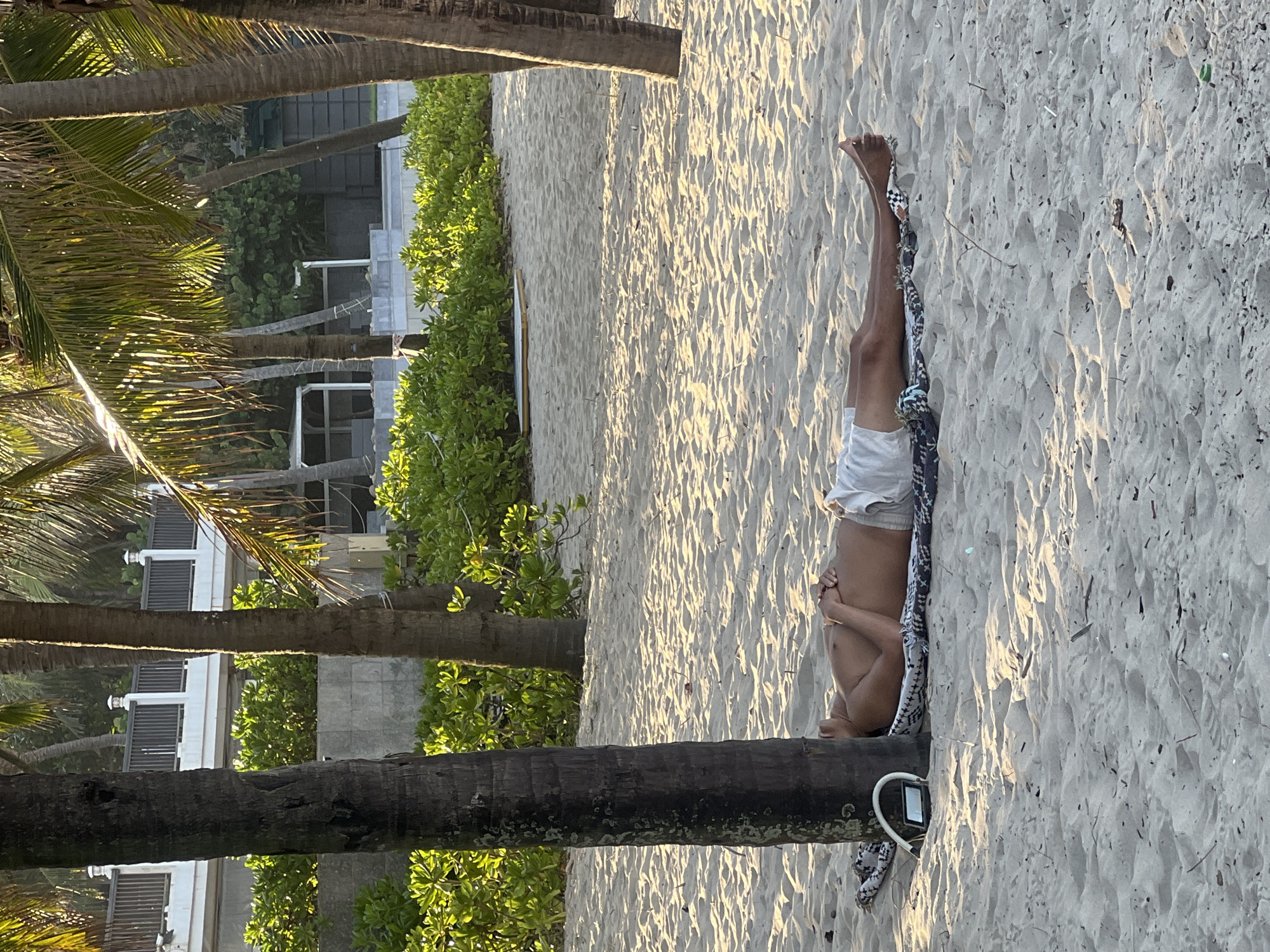 A person lies on their back on a sandy beach, resting on a patterned blanket. They are wearing white shorts and their legs are extended upwards. The scene is set on a beach in Da Nang, Vietnam, with palm trees framing the left side of the image. Sunlight casts long shadows across the sand, suggesting it is either early morning or late afternoon. Lush green foliage and modern buildings are visible in the background, indicating a developed coastal area. The person's posture and the relaxed setting suggest a moment of leisure and peace. No visible text is present in the image.