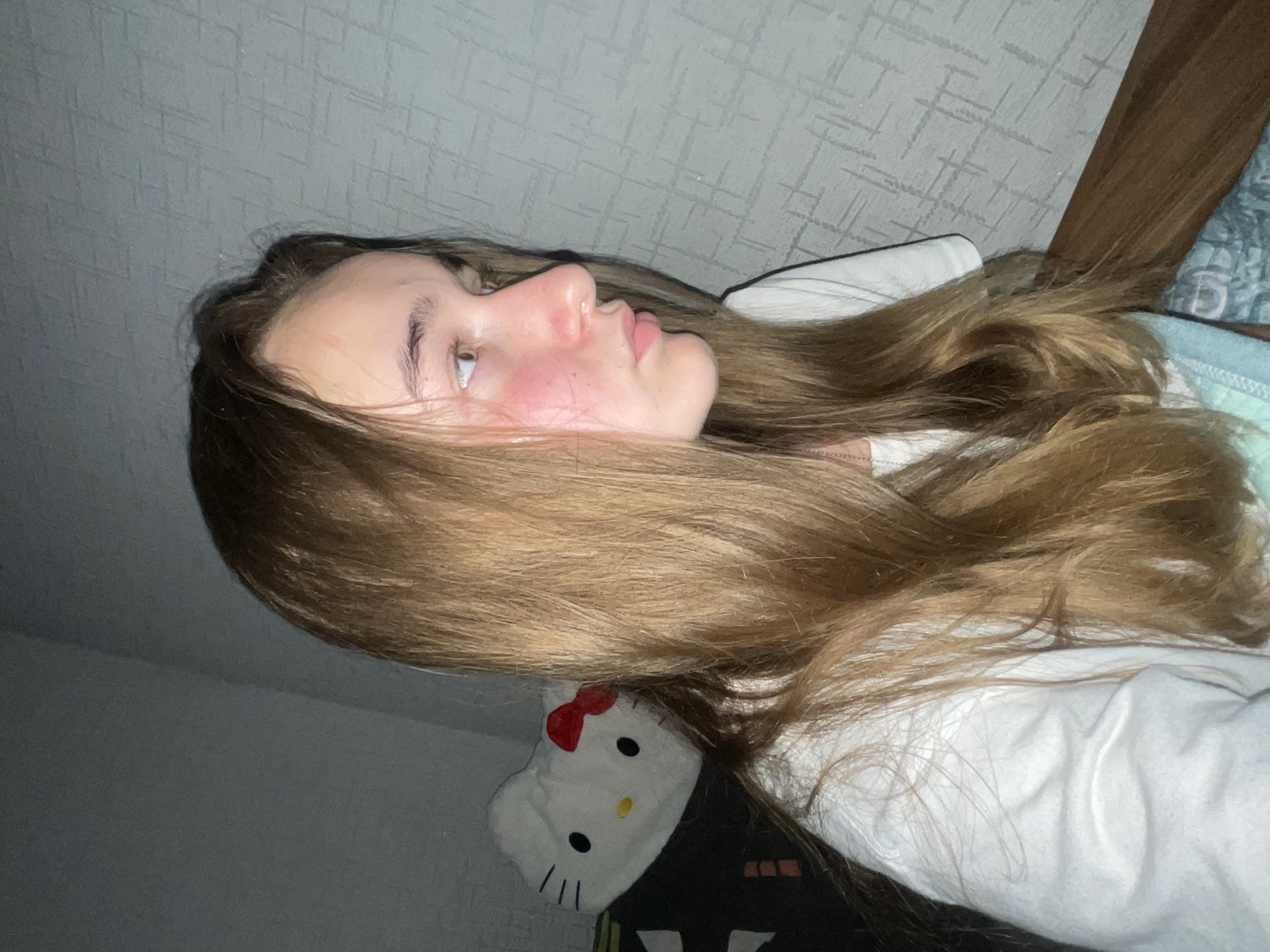 A medium shot photograph captures a young person with long, light brown hair, seen in profile from the right side of their face. They are wearing a white t-shirt and have noticeable red flushing on their cheeks. The background consists of a textured gray wall and a wooden surface along the bottom right of the frame. To the left, a white plush toy resembling Hello Kitty with a red bow is visible. The scene appears to be indoors, likely in a residential setting in Novopillia, Ukraine.