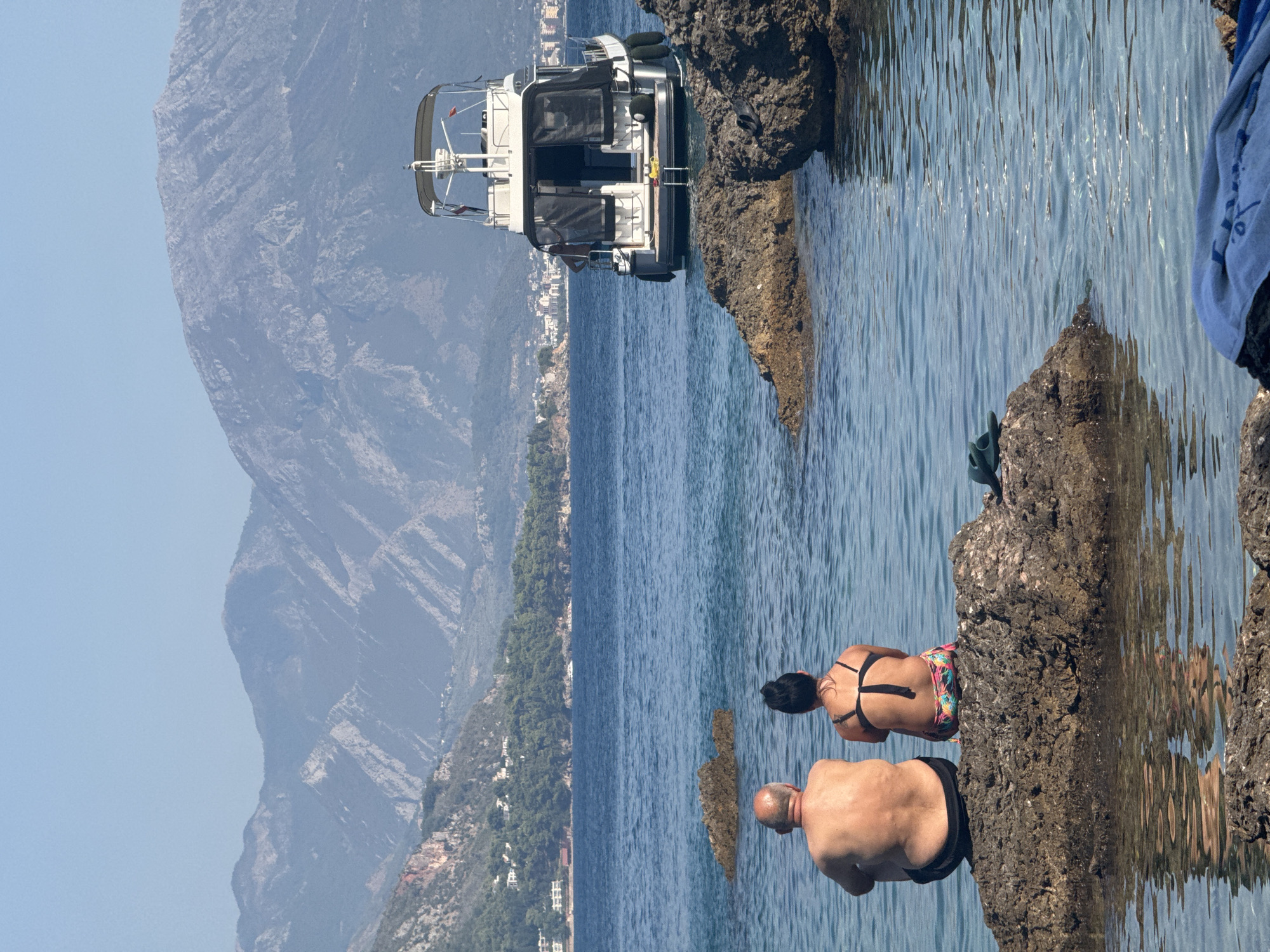 Two people, a man and a woman, sit on a large rock at the edge of the water, facing away from the camera. The man is bald and appears to be overweight. The woman has dark hair pulled back and is wearing a colorful bikini. They are seated on a large, rough rock that is partially submerged in clear, blue water. In the background, a white motor yacht is anchored offshore. Beyond the yacht, a coastline with buildings and lush green trees is visible, backed by a range of imposing, grey, rocky mountains under a clear blue sky. The scene appears to be a coastal area in or near Sutomore, Montenegro.