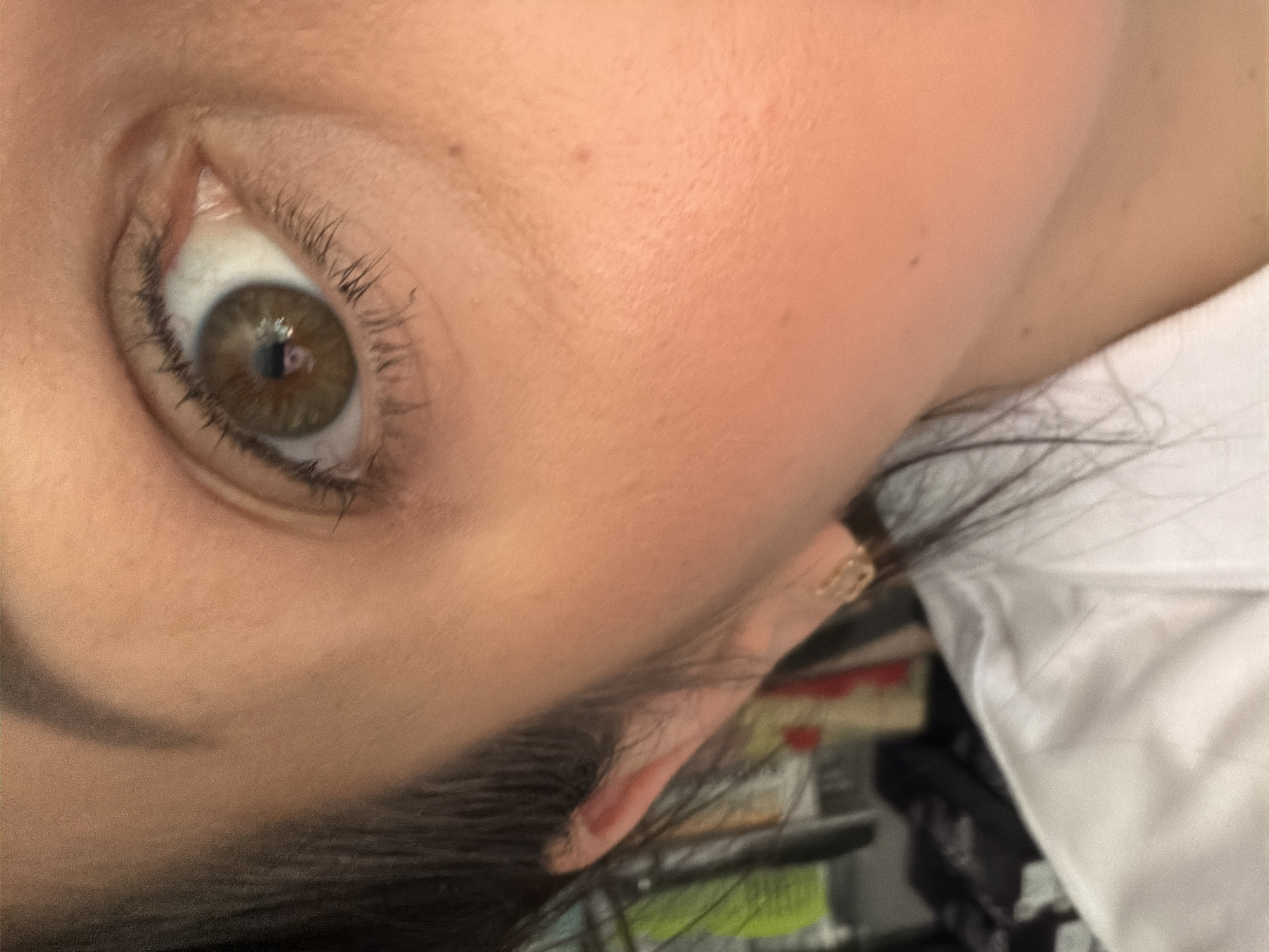 This close-up image, rotated counter-clockwise, features a person's left eye and a portion of their face. The eye has a striking hazel iris, displaying a mix of green and brown tones, with a clear reflection of a light source in the pupil. Long, dark eyelashes frame the eye, suggesting the application of mascara. The skin surrounding the eye and extending to the cheek appears smooth, exhibiting a fair to medium complexion with a few subtle blemishes or freckles. Dark hair is visible, framing the ear, where a small, gold-colored earring with a circular design adorns the earlobe. A white garment, likely a shirt or blouse, is partially visible in the bottom right corner of the frame. The background is softly blurred, indicating an indoor setting with indistinct objects, possibly furniture or shelves. The lighting appears to be artificial, highlighting the eye. The person's expression is neutral, and no specific activity is occurring beyond a direct gaze. There is no visible text in the image. While the provided location context is Sochi, Russia, no visual cues within the image confirm this.