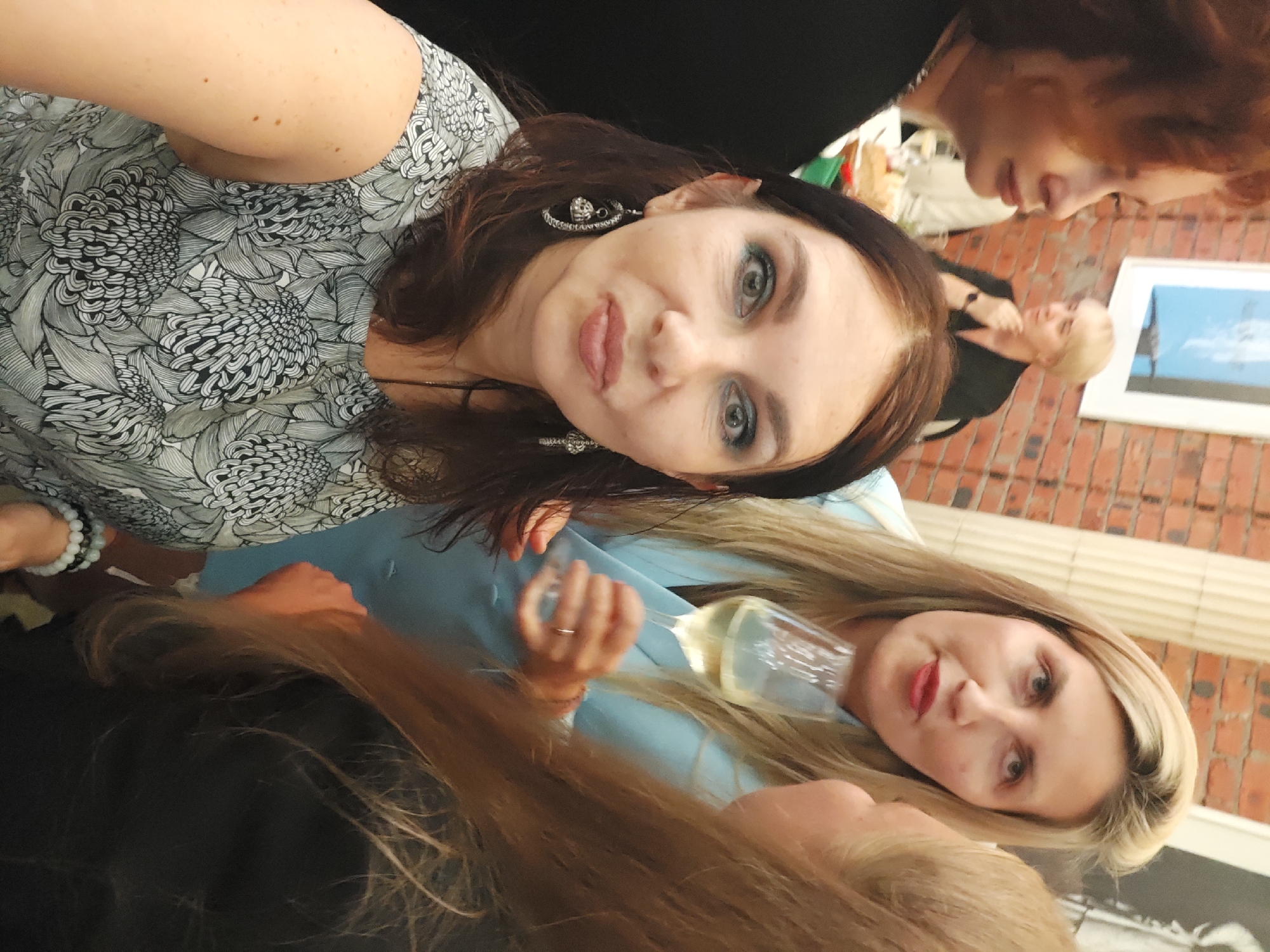 In Saint Petersburg, Russia, at a gathering, a woman with dark hair and prominent earrings is taking a selfie. She is wearing a grey and white floral print top. To her right, a blonde woman in a light blue blazer is holding a champagne glass. Behind them, another blonde woman with short hair is visible. The environment appears to be an indoor setting with brick walls and classical columns.