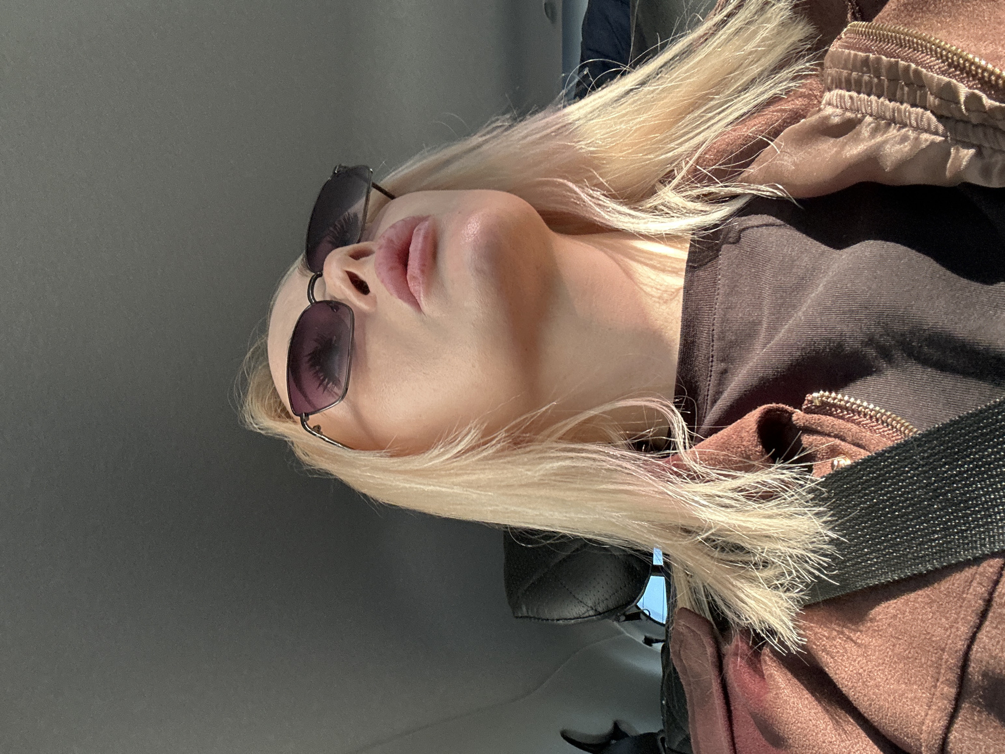 A woman with blonde hair and sunglasses is pictured in the interior of a car. She is wearing a dark top and a brown jacket with a seatbelt across her chest. The car interior is a muted grey color. The image appears to have been taken from a low angle. It is difficult to determine the location of Megion, Russia from the image.