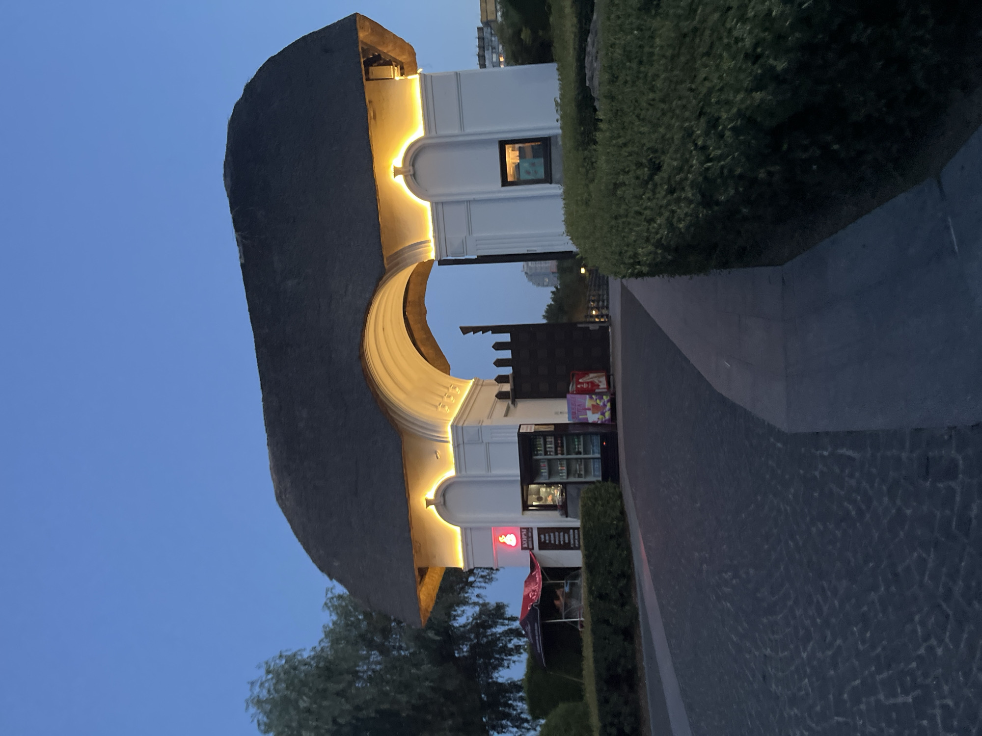An arched entrance with a thatched roof is illuminated by yellow lights against a twilight sky. The entrance features white pillars with arched niches. To the left of the entrance, there is a red umbrella over a stand, and a refrigerated display case filled with drinks. To the right, a tall, dark gate is visible. The ground is paved with cobblestones. Bushes line the sides of the pathway leading to the entrance. In the background, there are buildings and trees. The scene appears to be at the entrance to a park or attraction in Dnipro, Ukraine, likely at dusk.