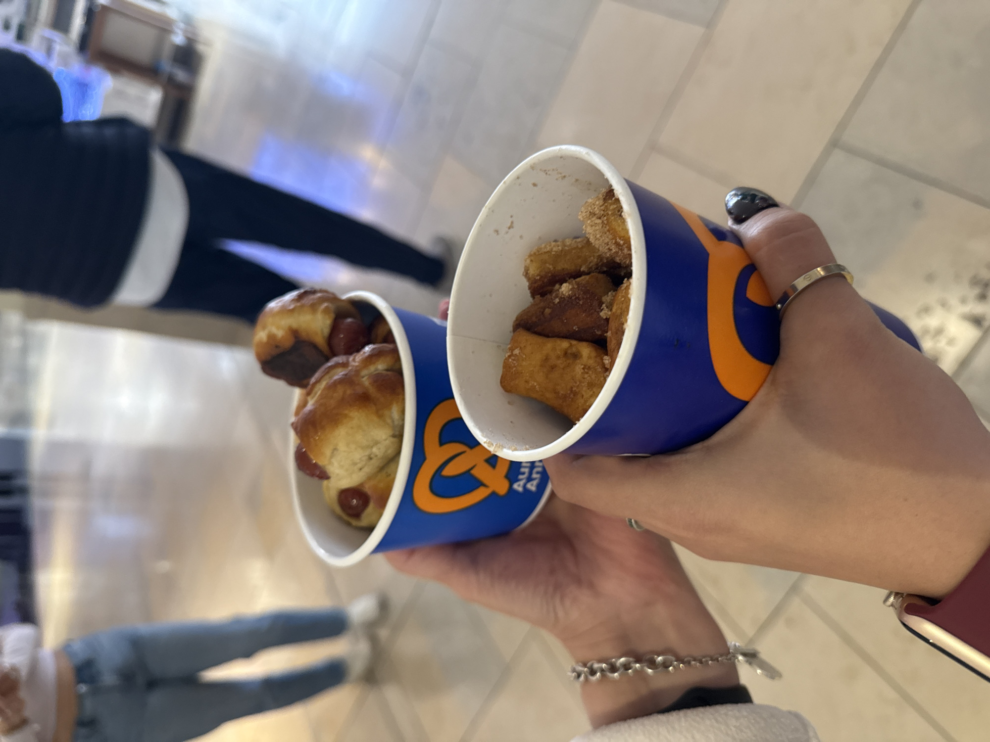A first-person view presents two cups of Auntie Anne's food being held indoors in an open area. The cup on the left contains pretzel dogs, while the cup on the right contains pretzel bites. The person holding the cups is wearing jewelry on their hands. In the background, two additional people are visible. The location is a mall in San Antonio, United States of America.